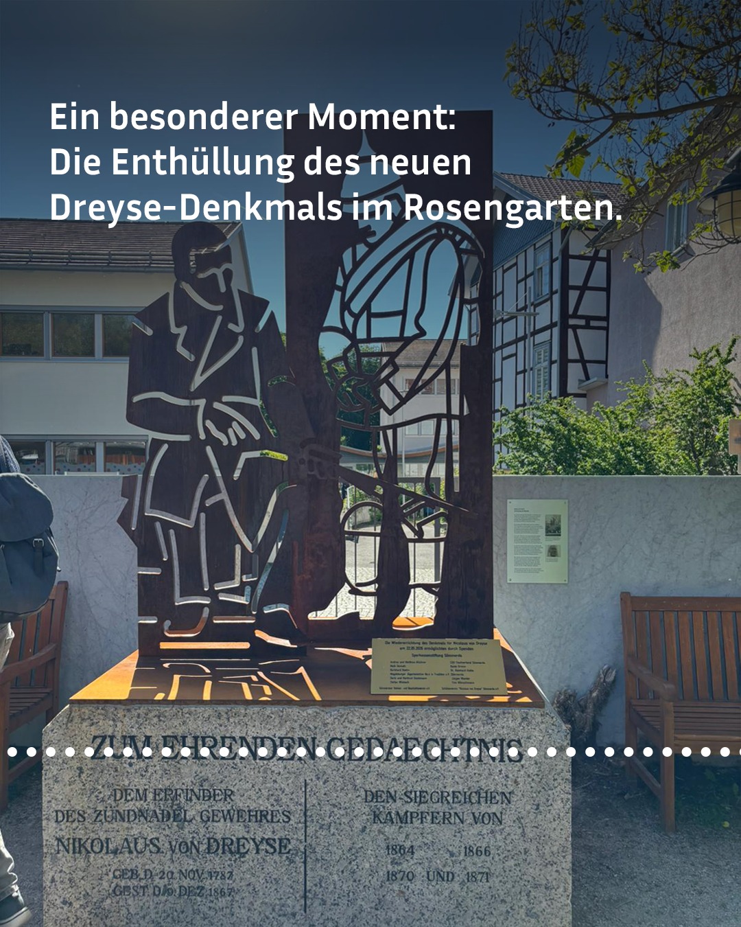 Metallskulptur des Nikolaus von Dreyse auf Granitsockel mit Inschrift, im Rosengarten vor Fachwerkhaus, mit Texteinblendung über die Enthüllung des Denkmals. (KI-Alternativtext)