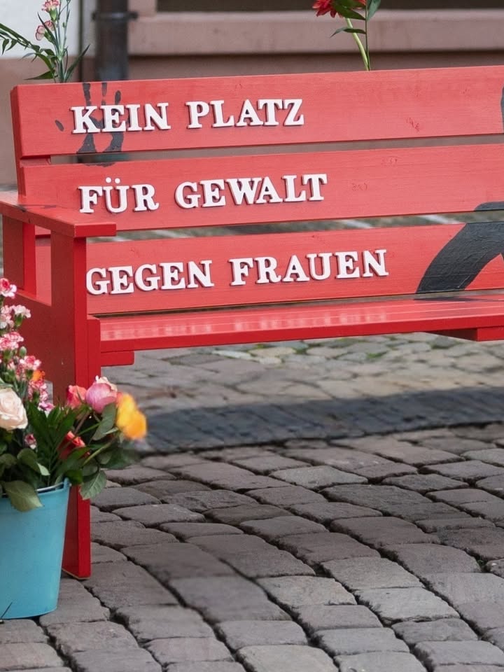 Eine rote Bank auf Kopfsteinpflaster trägt in weißen Buchstaben die Aufschrift „KEIN PLATZ FÜR GEWALT GEGEN FRAUEN“. Neben der Bank steht ein blauer Eimer mit Blumen, und auf der Bank liegen weitere Blumen. (KI-Alternativtext)