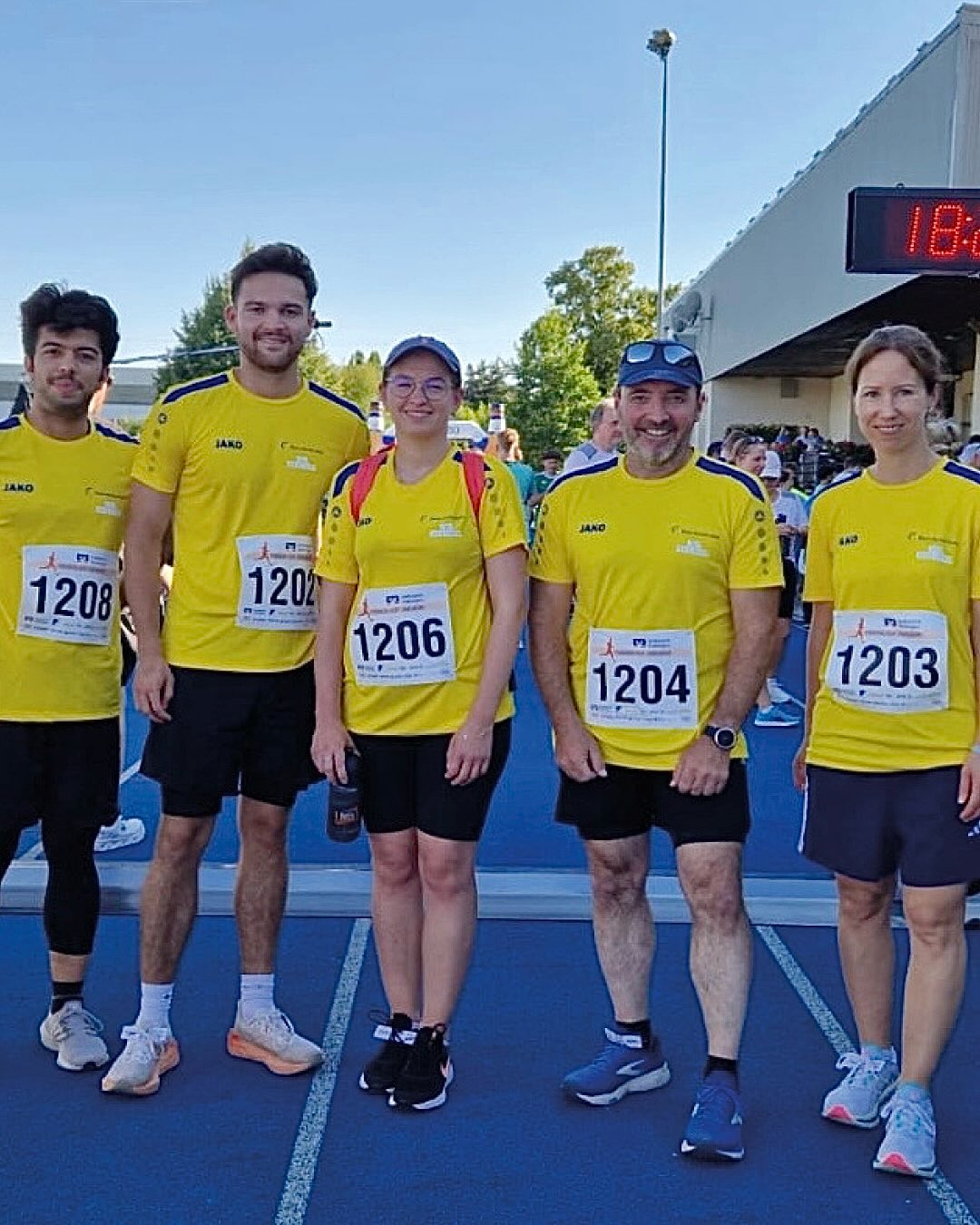 Fünf Personen in gelben T-Shirts und schwarzen Shorts stehen nebeneinander auf einer blauen Laufbahn. Sie tragen Startnummern, die von links nach rechts 1208, 1202, 1206, 1204 und 1203 lauten. Im Hintergrund ist eine digitale Uhr zu sehen. (KI-Alternativtext)