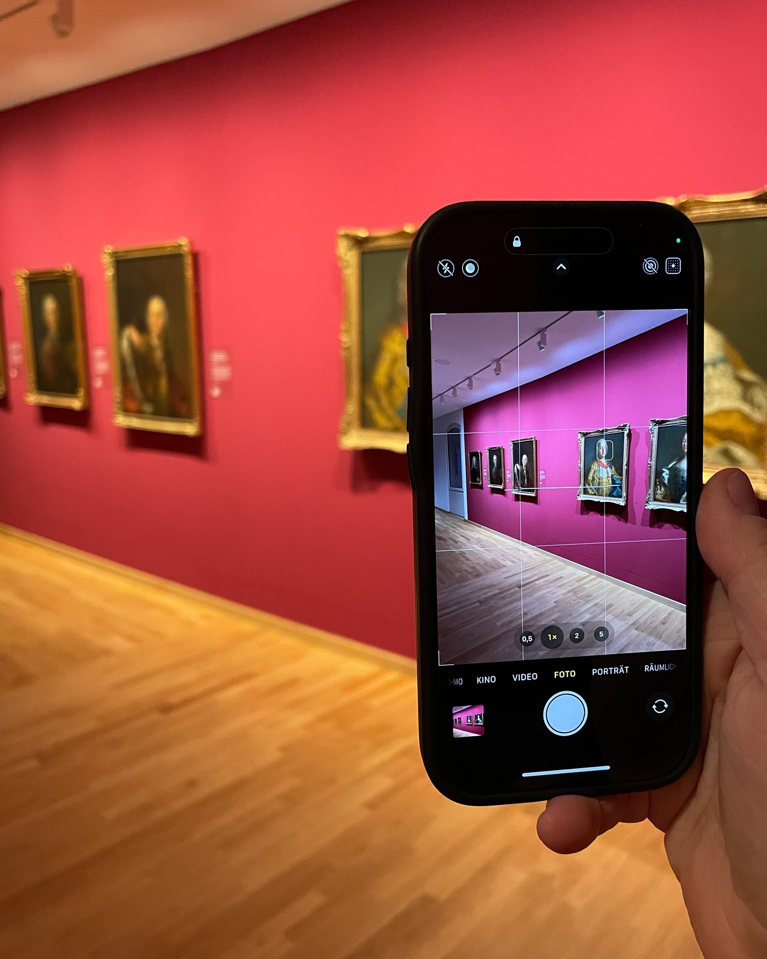 Eine Hand hält ein Smartphone, das im Foto-Modus eine Galerie mit roten Wänden und gerahmten Porträts aufnimmt; im Display sind die Kameraeinstellungen und ein Raster sichtbar. (KI-Alternativtext)