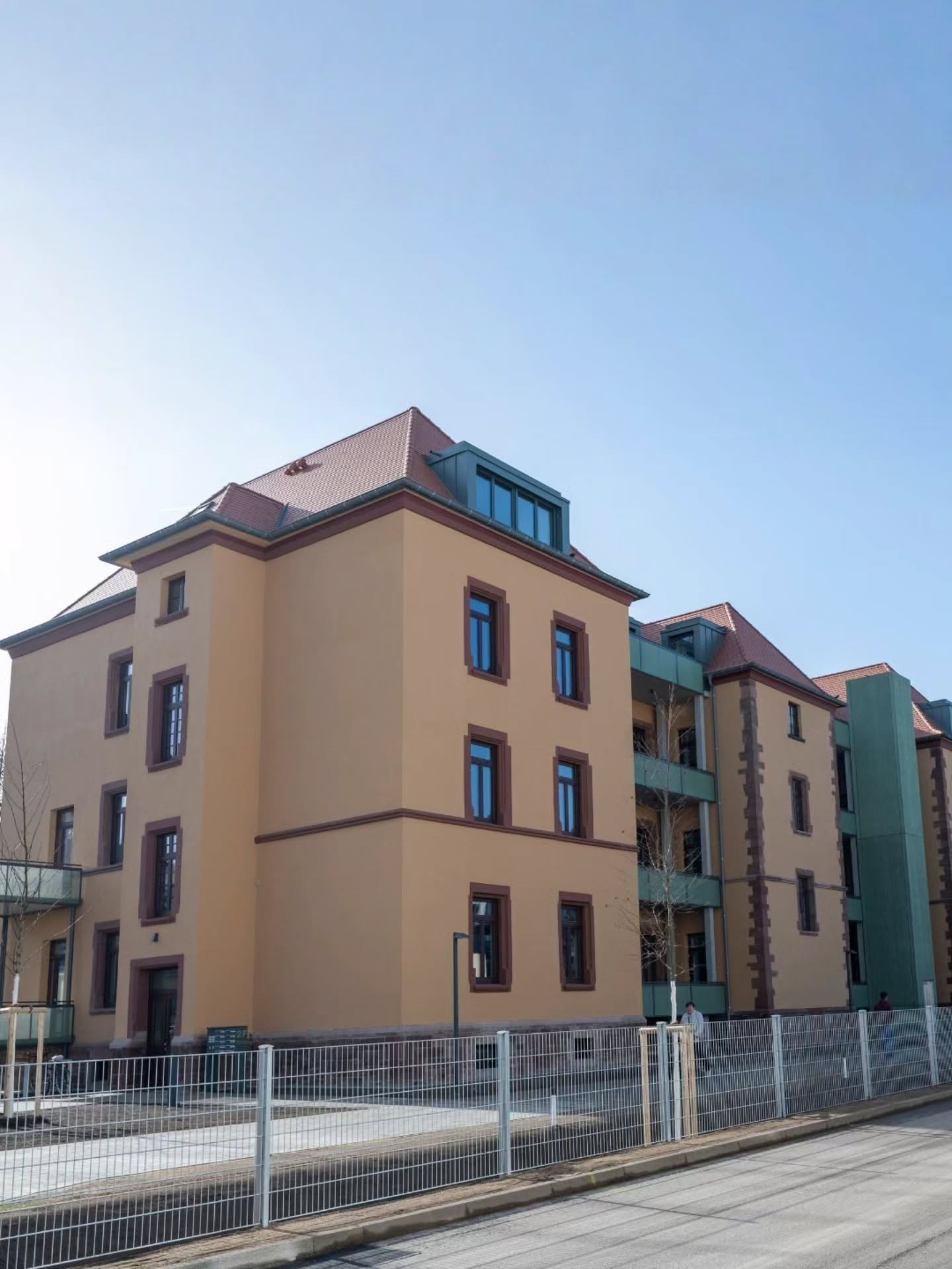 Dieser Alternativtext wurde KI-generiert: Ein Gebäude mit einem roten Ziegeldach und einer hellen Fassade steht vor blauem Himmel. Ein Zaun begrenzt das Grundstück vor dem Gebäude.