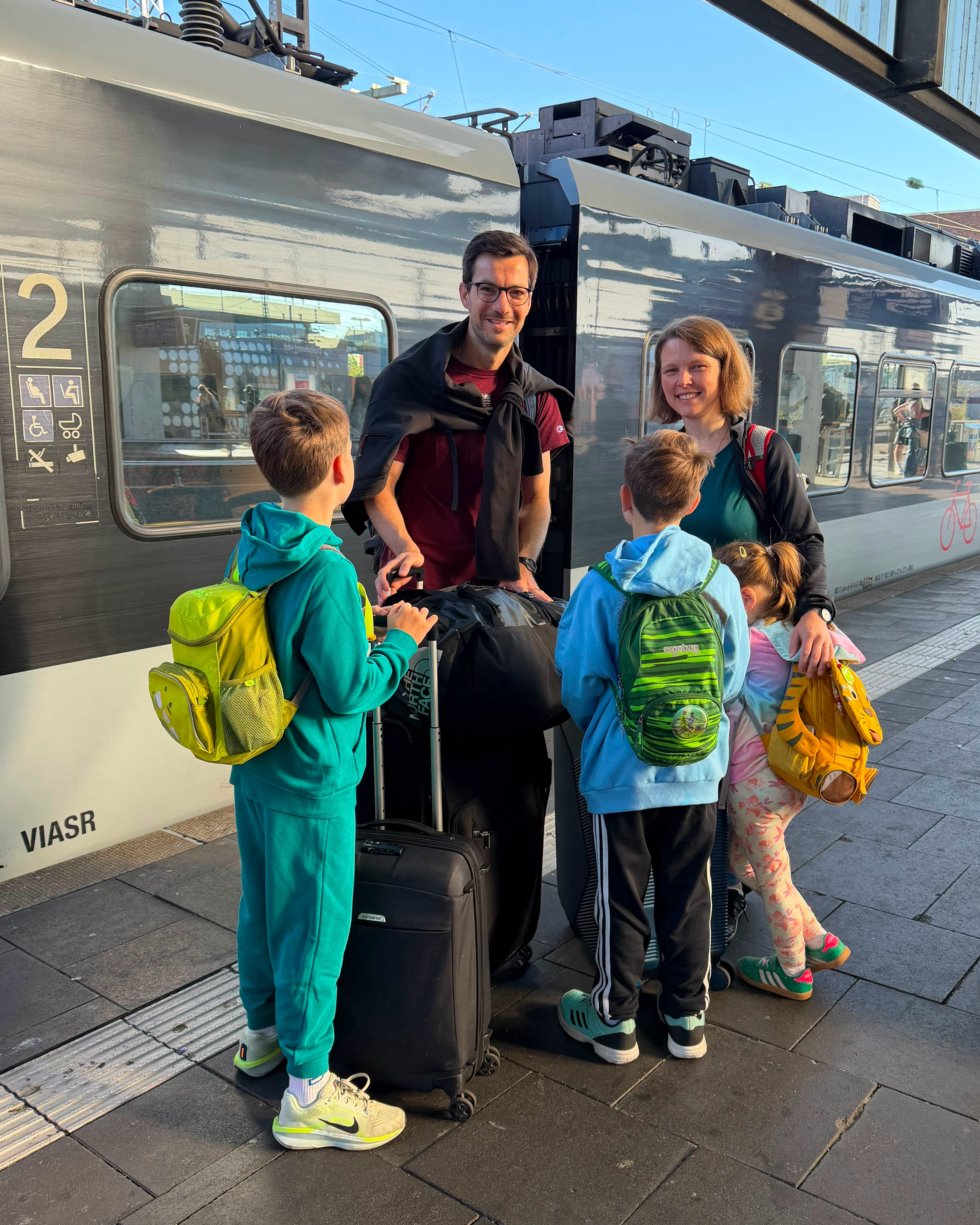 Eine Gruppe von Erwachsenen und Kindern steht auf einem Bahnsteig neben einem Zug mit der Aufschrift "VIASR" und der Zahl "2". Sie haben Koffer und Rucksäcke dabei und lächeln in die Kamera. (KI-Alternativtext)