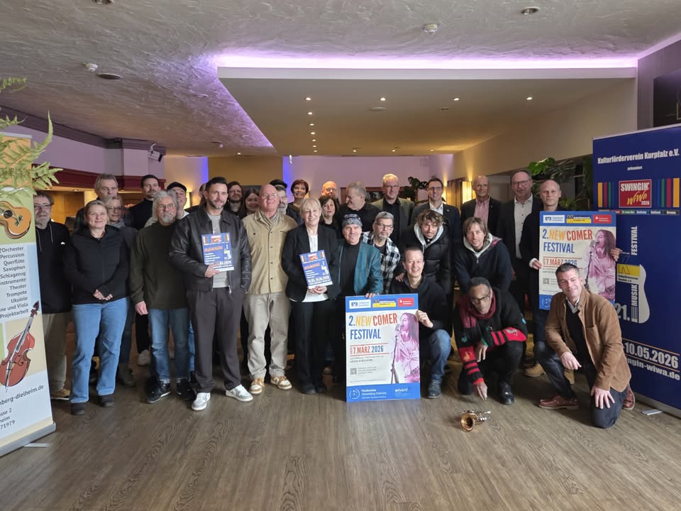 Dieser Alternativtext wurde KI-generiert: Eine Gruppe von Menschen steht in einem Raum zusammen und hält Poster. Im Hintergrund befindet sich eine Bühne und Werbematerialien für ein Musikfestival.