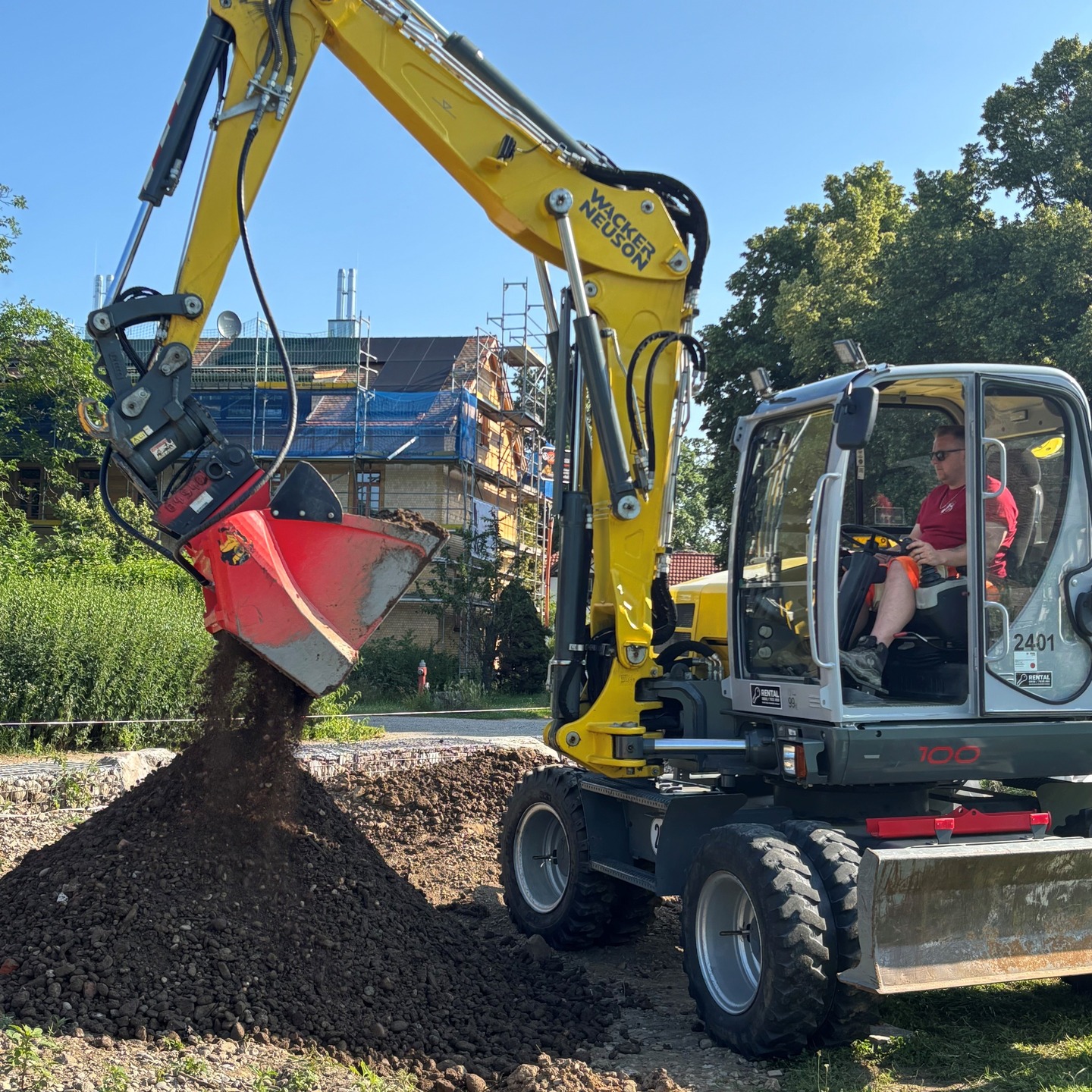 Ein gelber Bagger schaufelt Erde auf einen Haufen. Im Hintergrund befindet sich ein Gebäude mit Baugerüst.