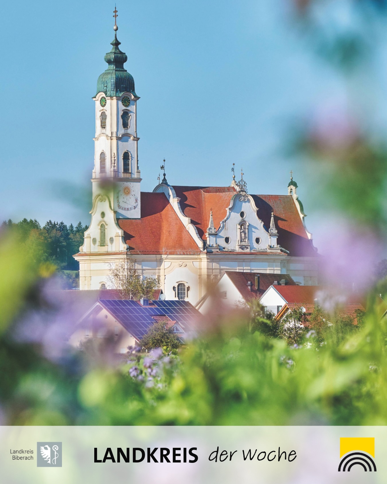 Barocke Kirche mit hohem Turm und Zwiebelhaube, umgeben von Häusern mit Solaranlagen, im Vordergrund unscharfe Blumen. Unten Text „LANDKREIS der Woche“ mit Logo des Landkreises Biberach. (KI-Alternativtext)