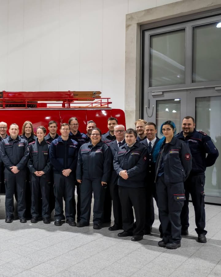 Dieser Alternativtext wurde KI-generiert: Eine Gruppe von Personen in Uniformen steht für ein Foto vor einer weißen Wand und einer Glasfassade. Im Hintergrund ist ein rotes Feuerwehrfahrzeug zu sehen.