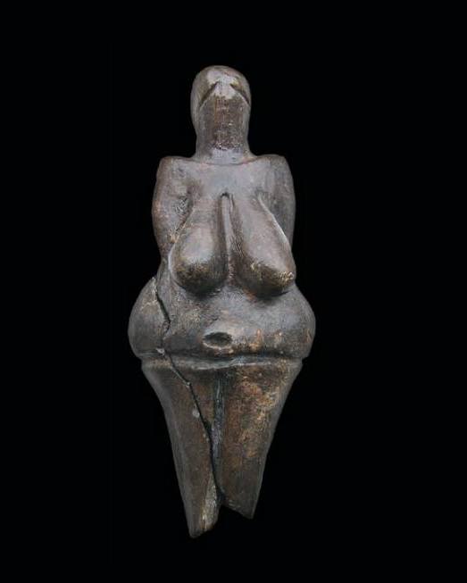 Eine kleine, bräunliche Steinfigur zeigt eine menschliche Gestalt mit stark betonten Merkmalen wie großen Brüsten, breiten Hüften und einem runden Bauch. Der Kopf ist von einer massigen Kopfbedeckung umgeben, während die Beine kurz sind und spitz zulaufen. (KI-Alternativtext)