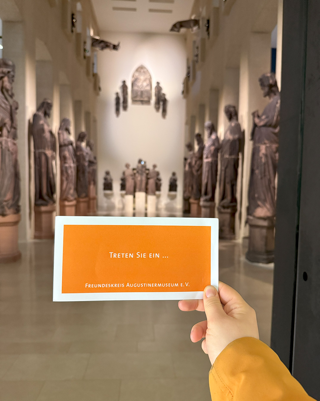 Dieser Alternativtext wurde KI-generiert: Ein Museumsbesucher hält einen orangefarbenen Flyer vor einer Galerie mit Skulpturen. Im Hintergrund sind weitere Figuren und ein Fenster zu sehen.