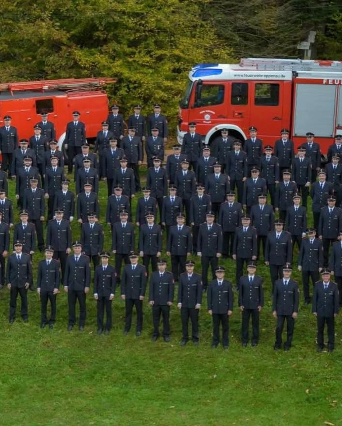 Eine große Gruppe von Personen in Uniformen steht auf einem Feld vor zwei roten Feuerwehrfahrzeugen.