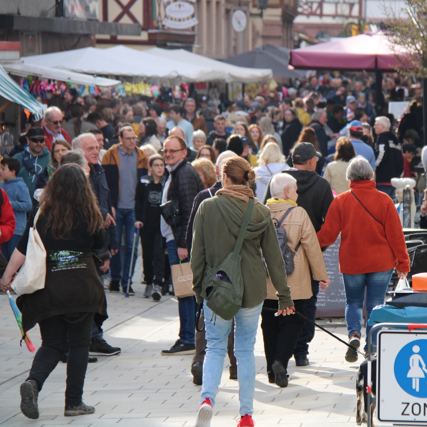 Menschen bewegen sich auf einem belebten Marktplatz mit Marktständen. Ein Hund ist an einer Leine zu sehen.