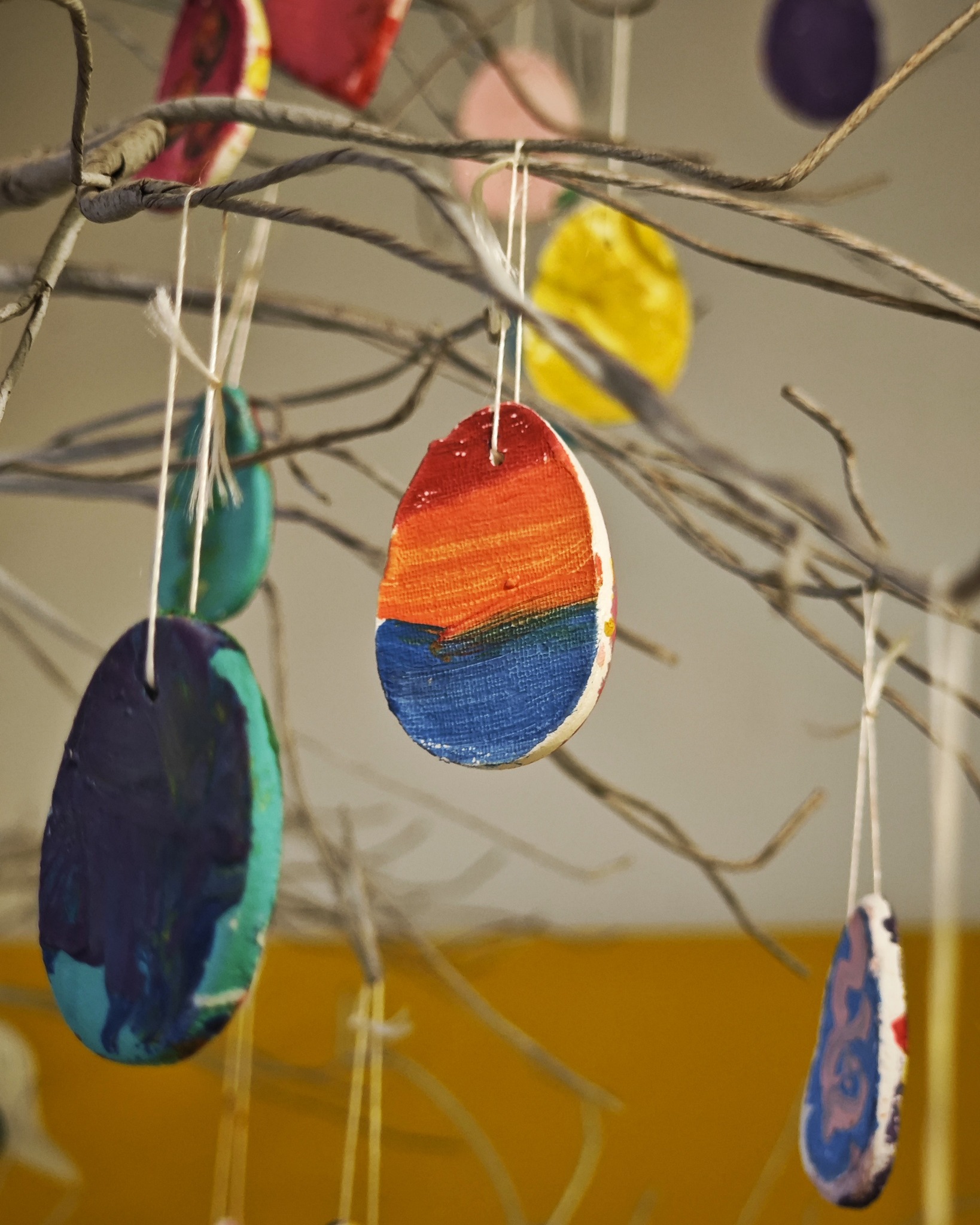 Dieser Alternativtext wurde KI-generiert: Bunte, bemalte Dekorationen aus einem festen Material hängen an dünnen Fäden von Ästen. Im Hintergrund ist eine gelbe Wand zu sehen.