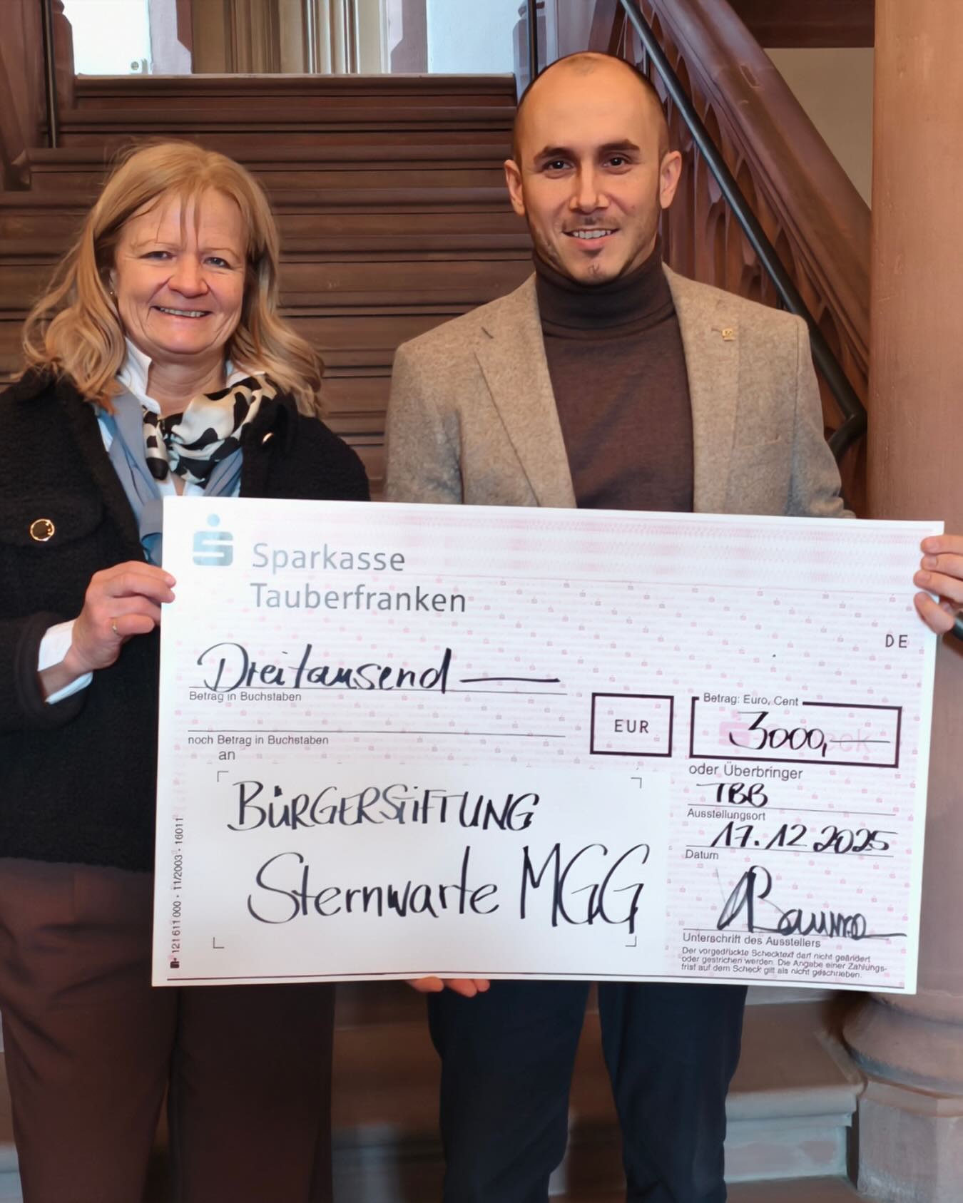 Dieser Alternativtext wurde KI-generiert: Eine Person überreicht einen großen Scheck an eine andere. Der Scheck ist für eine Spende an eine Bürgerstiftung ausgestellt.