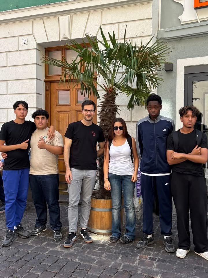 Sechs Personen stehen nebeneinander auf einem gepflasterten Boden vor einem Gebäude mit einer Holztür und einer Palme in einem Kübel. Die Gruppe trägt legere Kleidung, wobei sich zwei Personen auf der linken Seite die Arme um die Schultern gelegt haben und die Person ganz rechts die Arme verschränkt hält. Über der Tür ist die Hausnummer 2 angebracht. (KI-Alternativtext)