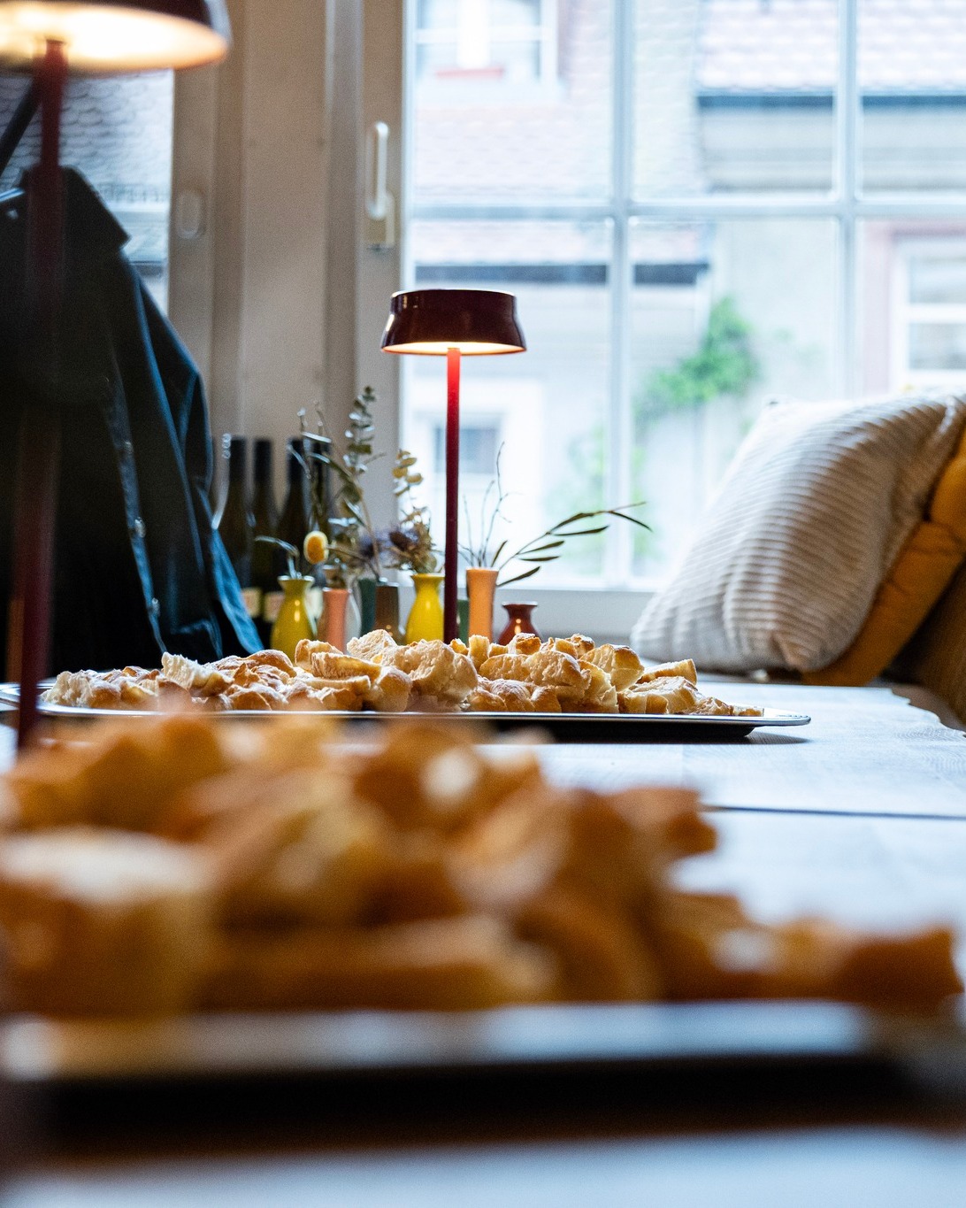 Auf einem Tisch stehen zwei Tabletts mit frisch gebackenen, goldbraunen Brötchen. Im Hintergrund leuchtet eine rote Tischlampe, daneben ein Fenster und ein Sofa mit Kissen. (KI-Alternativtext)