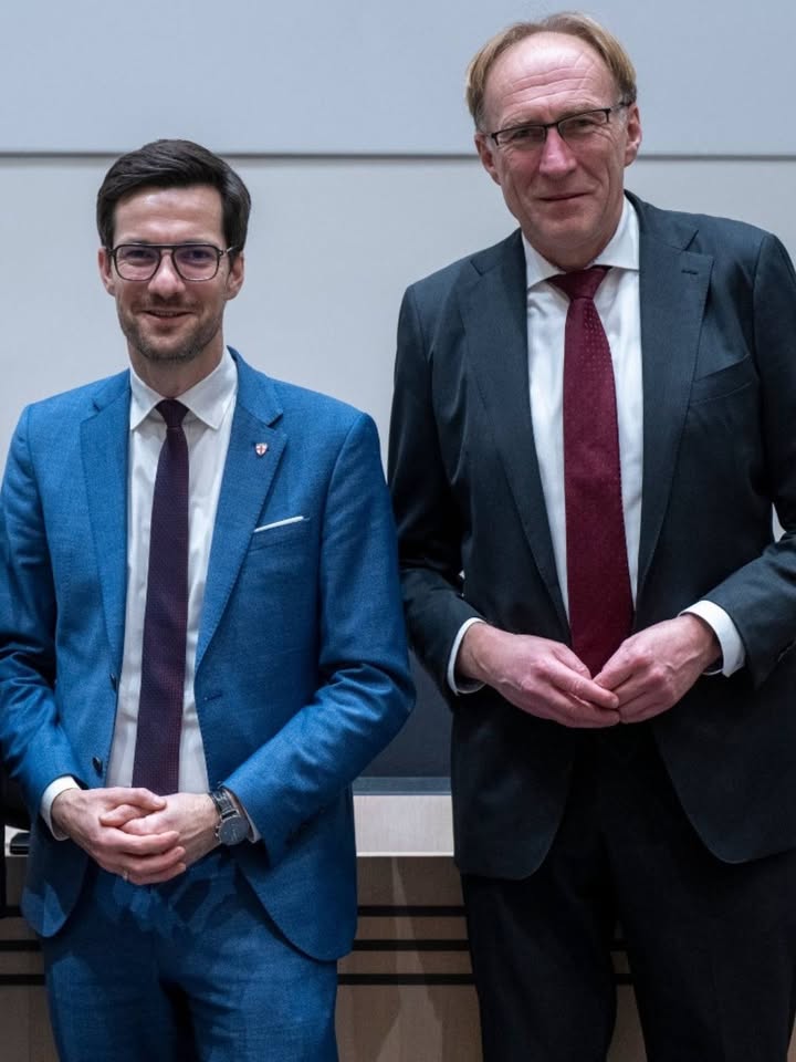 Dieser Alternativtext wurde KI-generiert: Zwei Personen in Anzügen stehen nebeneinander und lächeln in die Kamera. Im Hintergrund ist eine Wand mit einer Holzstruktur zu sehen.