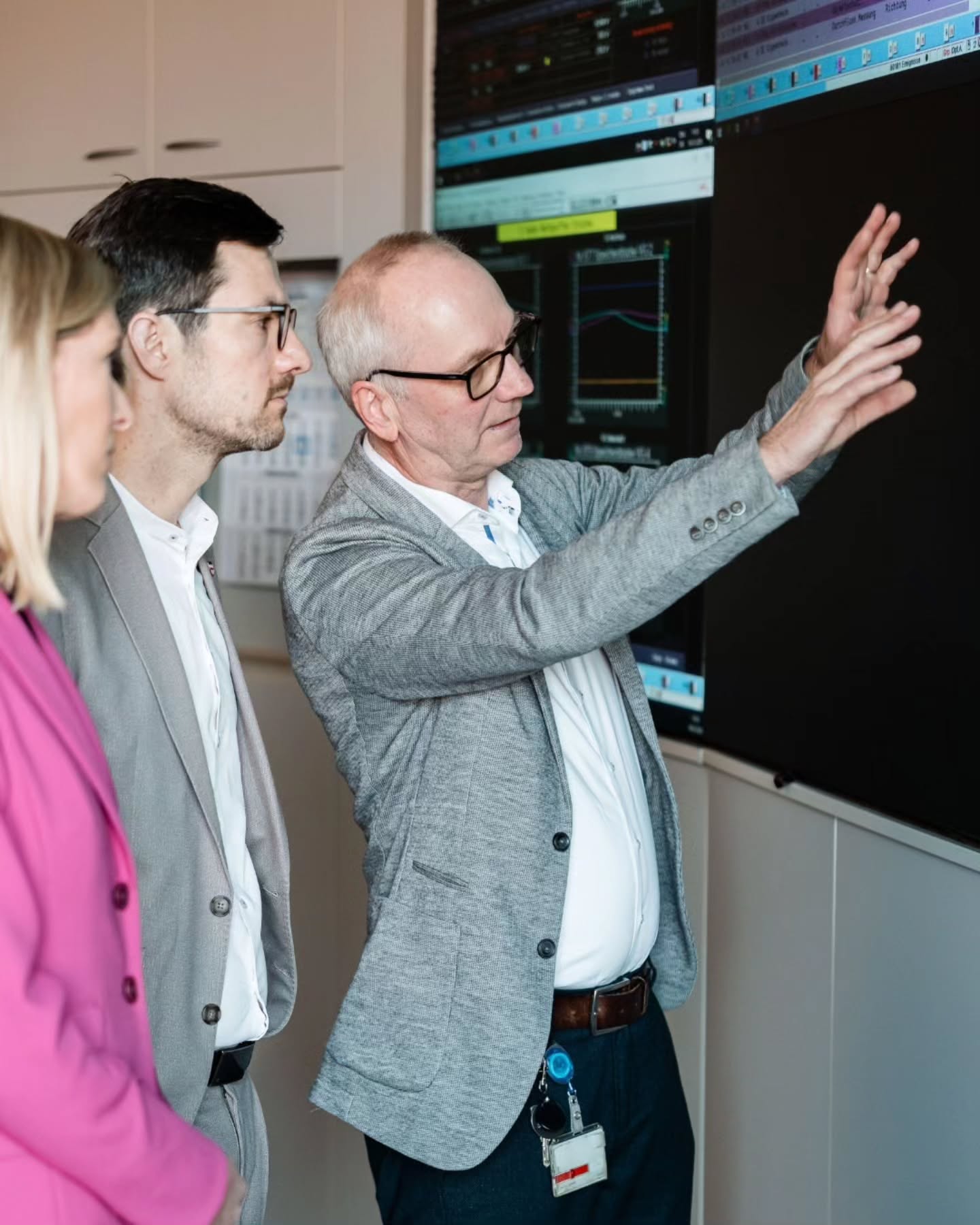 Dieser Alternativtext wurde KI-generiert: Zwei Personen betrachten einen Bildschirm und ein Diagramm an einer Wand. Eine Person deutet auf das Diagramm, während die andere Person zusieht.