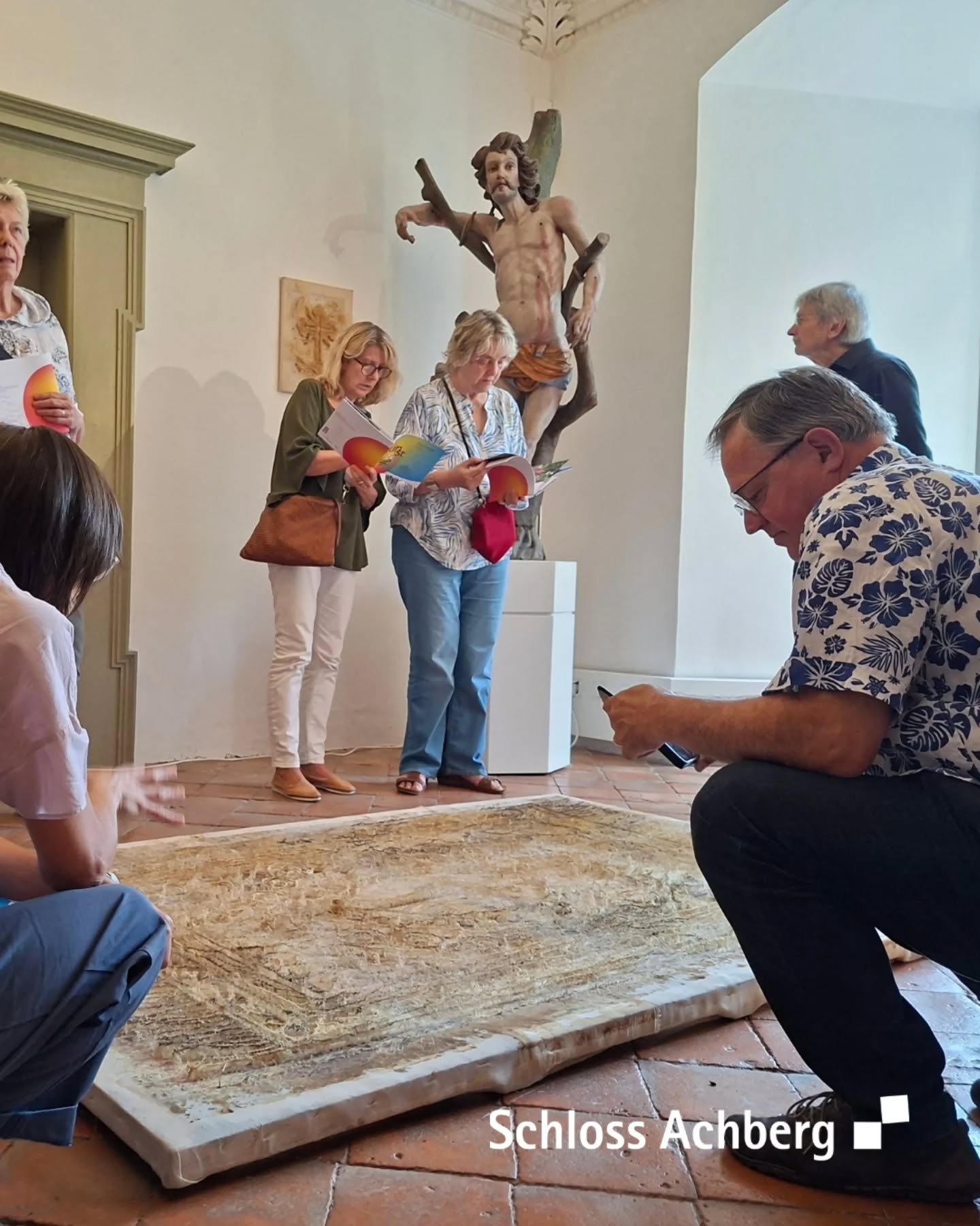Eine Gruppe betrachtet ein Kunstwerk in einem Raum. Auf einem Sockel steht zudem eine Skulptur.