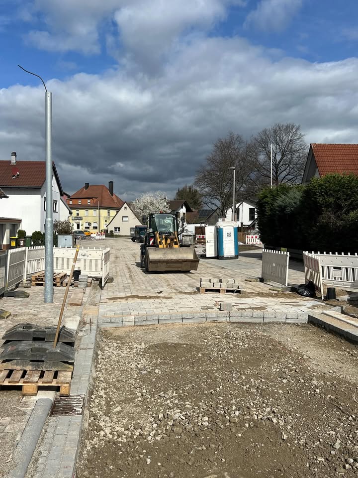 Dieser Alternativtext wurde KI-generiert: Ein Straßenbau ist im Gange. Im Vordergrund sind Baumaterialien und ein Bagger zu sehen, im Hintergrund Wohnhäuser und eine bewölkte Landschaft.