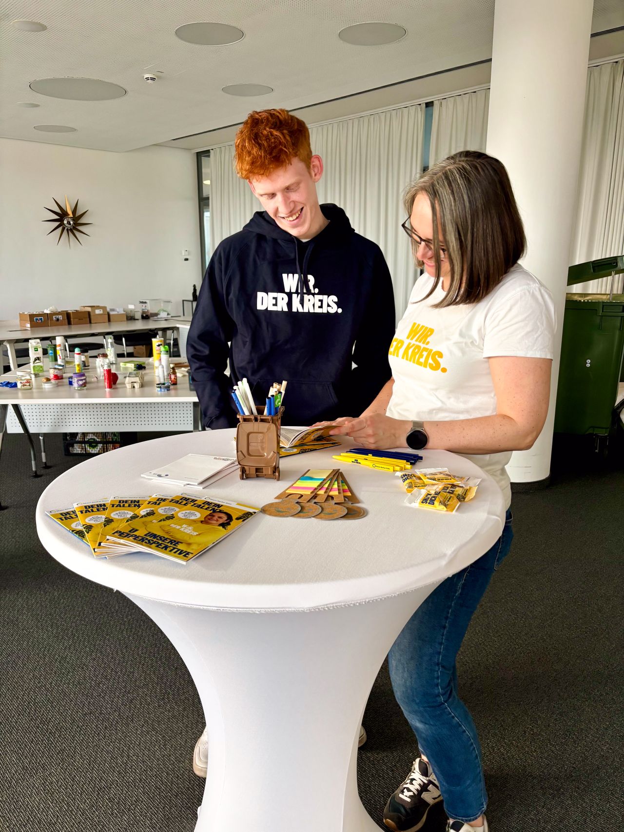 Dieser Alternativtext wurde KI-generiert: Zwei Personen stehen an einem Tisch mit Informationsmaterial. Im Hintergrund sind weitere Gegenstände und eine helle Büroatmosphäre zu sehen.