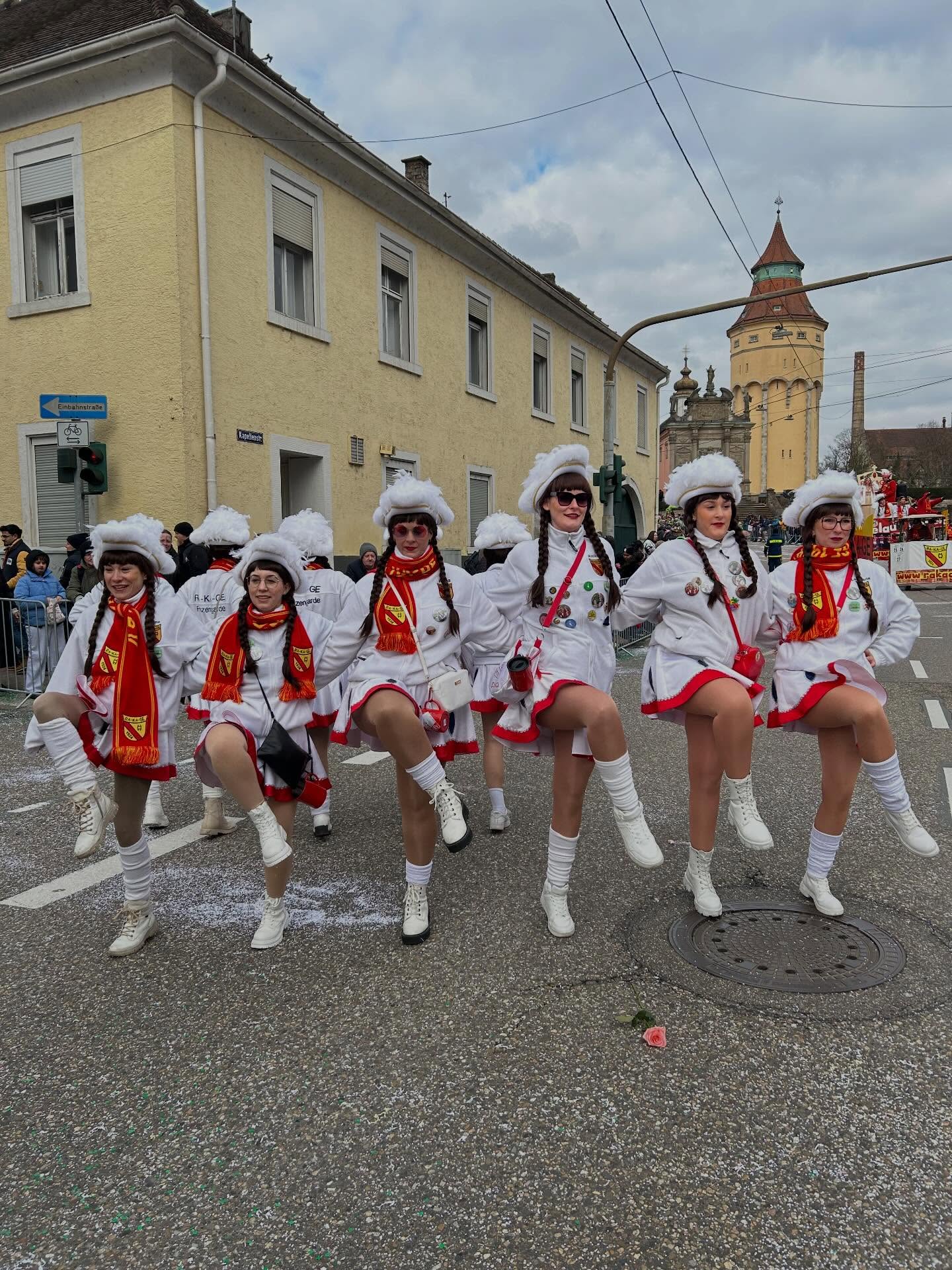 Dieser Alternativtext wurde KI-generiert: Eine Gruppe von Personen in traditioneller Tracht marschiert auf einer Straße. Im Hintergrund sind Gebäude und weitere Menschen zu sehen.