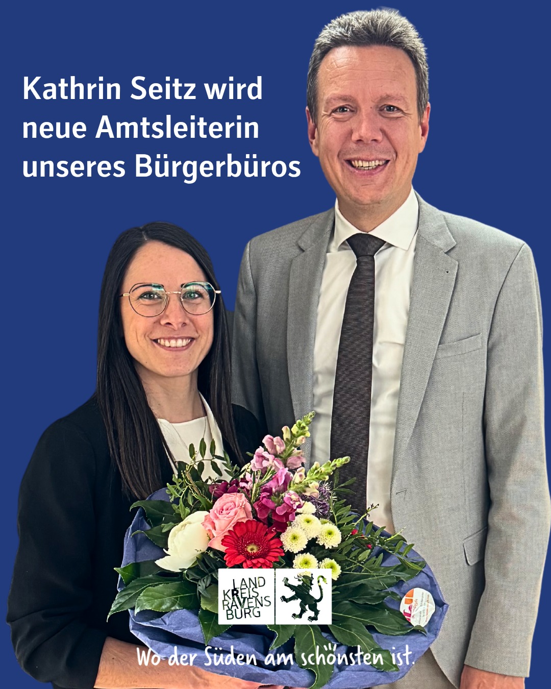 Zwei lächelnde Personen posieren vor einem blauen Hintergrund, wobei eine Person einen Blumenstrauß hält. Die Person im Anzug steht dahinter.