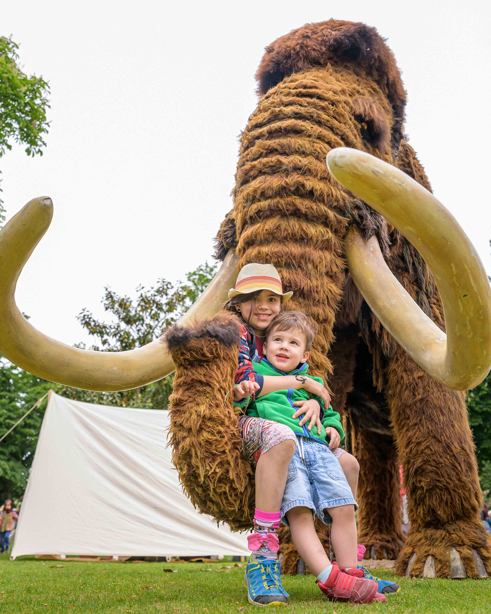 Zwei Kinder sitzen lächelnd auf dem Stoßzahn einer riesigen, pelzigen Mammut-Statue im Freien. Im Hintergrund sind Bäume und ein weißes Zelt zu sehen. (KI-Alternativtext)