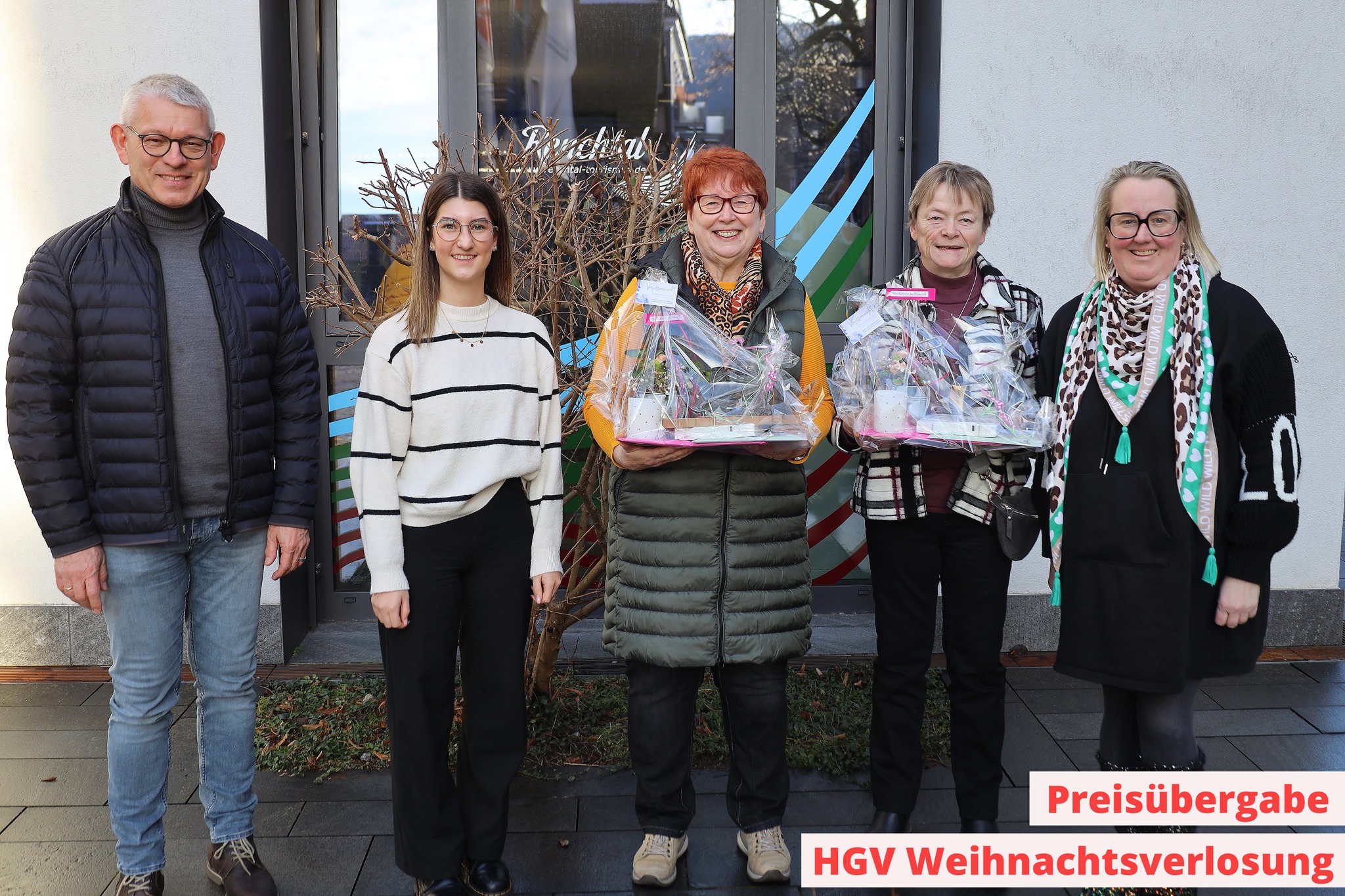 Dieser Alternativtext wurde KI-generiert: Eine Gruppe von Menschen steht vor einem Geschäft und hält verpackte Gegenstände. Ein Schild weist auf eine Preisübergabe im Rahmen einer Weihnachtsverlosung hin.