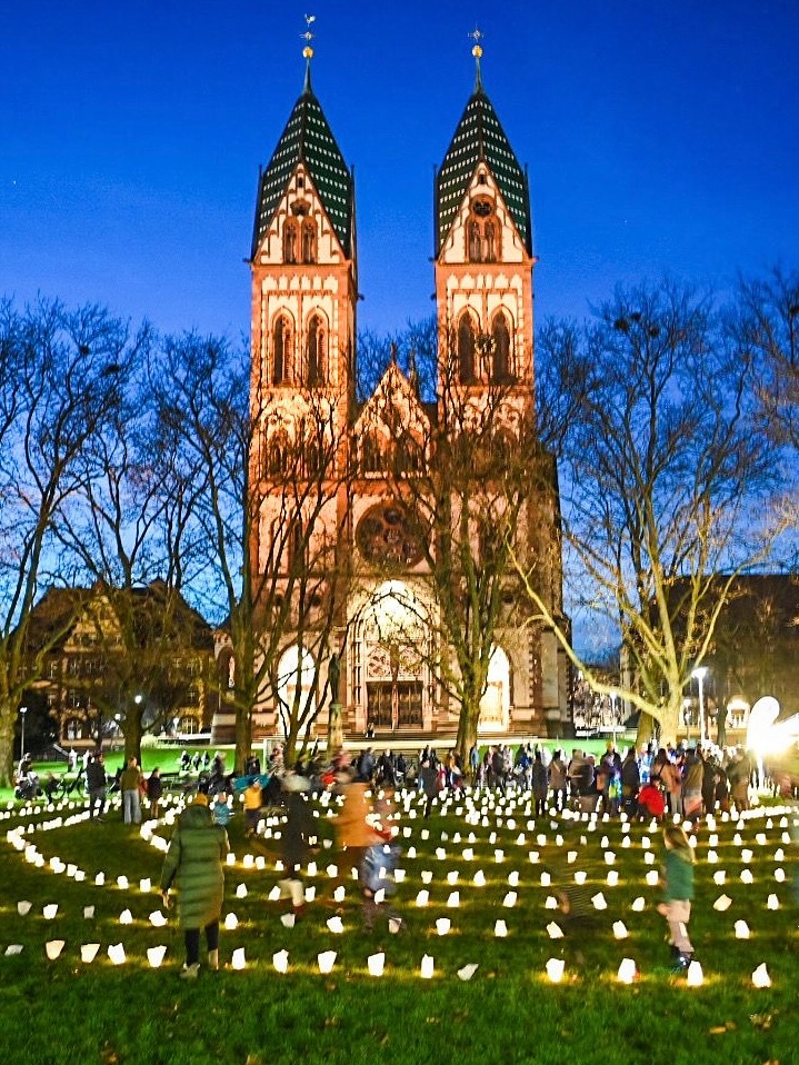 Dieser Alternativtext wurde KI-generiert: Ein großes, historisches Gebäude mit zwei Türmen ist bei Nacht beleuchtet. Im Vordergrund befinden sich viele kleine, leuchtende Kerzen auf einer Wiese und einige Menschen.