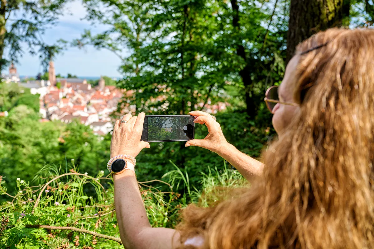 Eine Person mit langen, lockigen Haaren und Sonnenbrille macht mit einem Smartphone ein Foto einer Stadtansicht, die von Bäumen umgeben ist. Auf dem Handgelenk trägt die Person eine Smartwatch. (KI-Alternativtext)