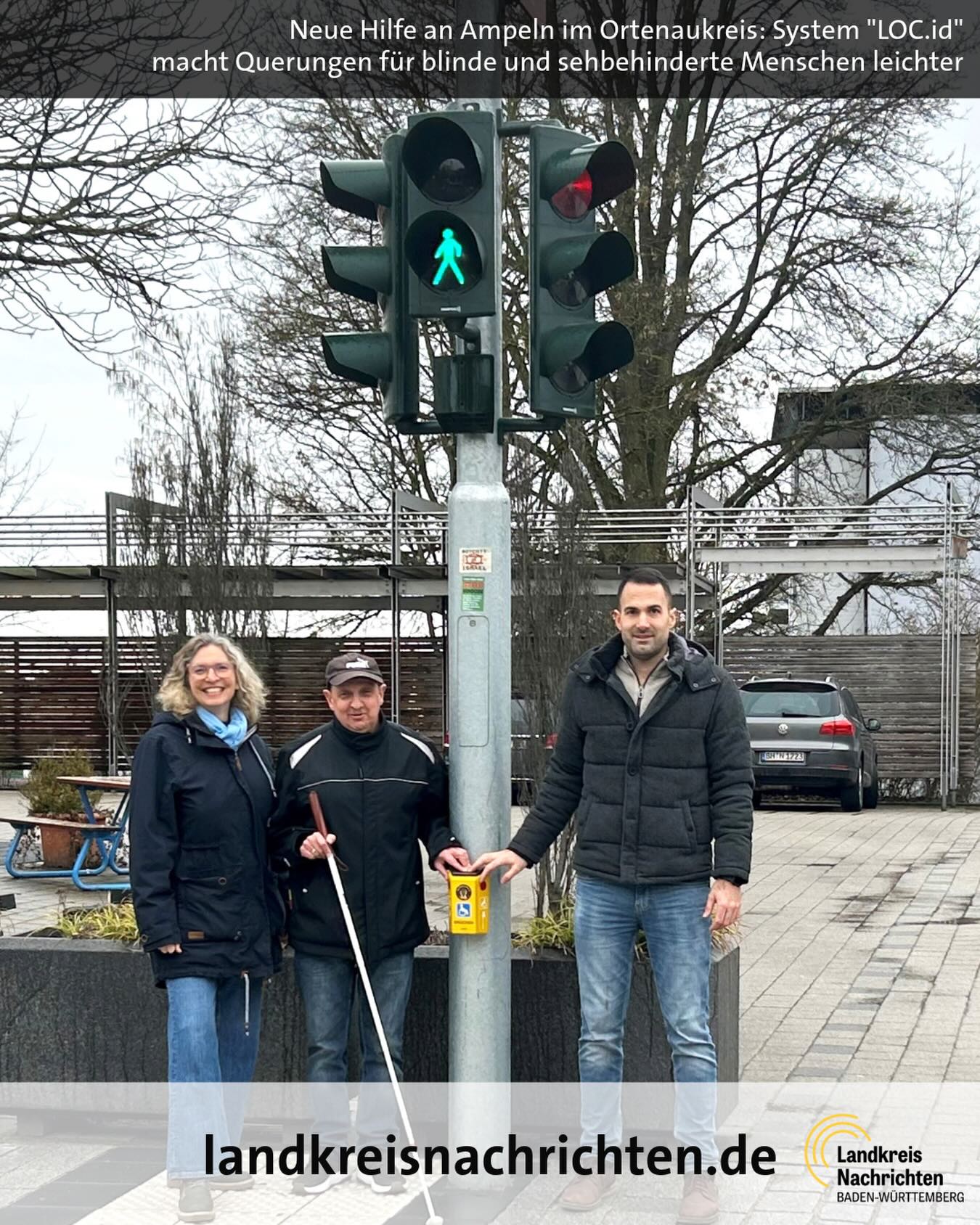 Dieser Alternativtext wurde KI-generiert: Eine Ampel mit akustischem Signalgerät steht im Vordergrund. Mehrere Personen stehen davor und blicken in die Kamera.