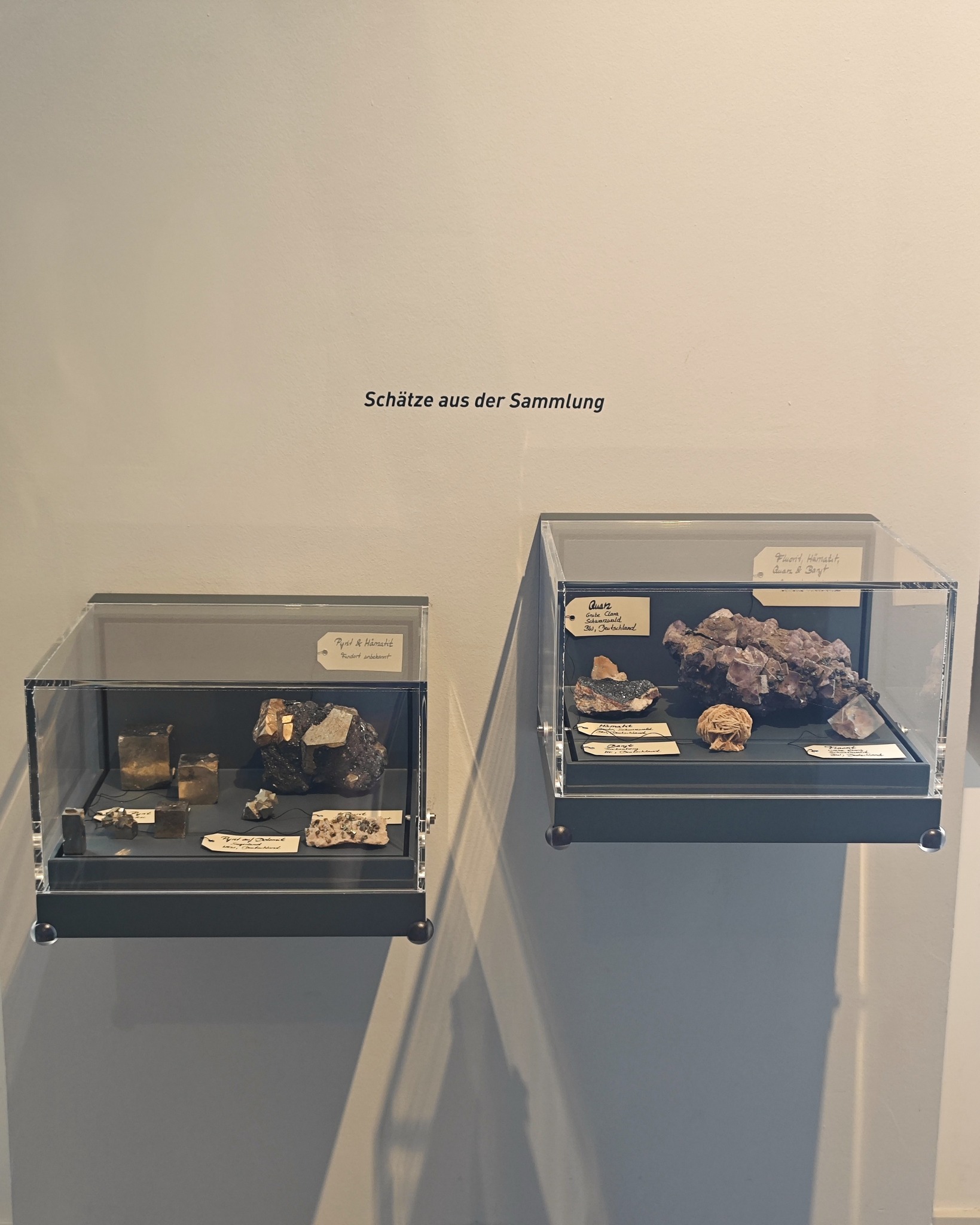 Zwei transparente Vitrinen hängen an einer Wand unter der Aufschrift „Schätze aus der Sammlung“. Die linke Vitrine enthält dunkle Mineralproben und Kristalle mit Etiketten wie „Pyrit & Hämatit“. Die rechte Vitrine zeigt größere Mineralaggregate, darunter ein großes violettes Kristallgebilde und eine kugelförmige Probe, ebenfalls mit Beschriftungen versehen. (KI-Alternativtext)