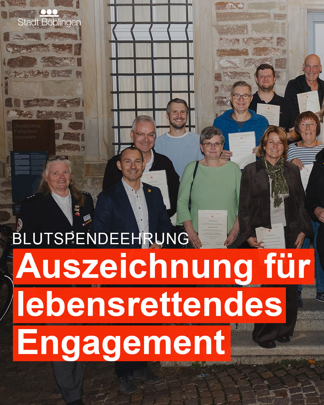 Dieser Alternativtext wurde KI-generiert: Eine Gruppe von Menschen steht lächelnd vor einem historischen Gebäude mit einem Banner, auf dem eine Auszeichnung für lebensrettendes Engagement zu sehen ist. Das Bild ist farbig und zeigt eine Zeremonie im Freien.