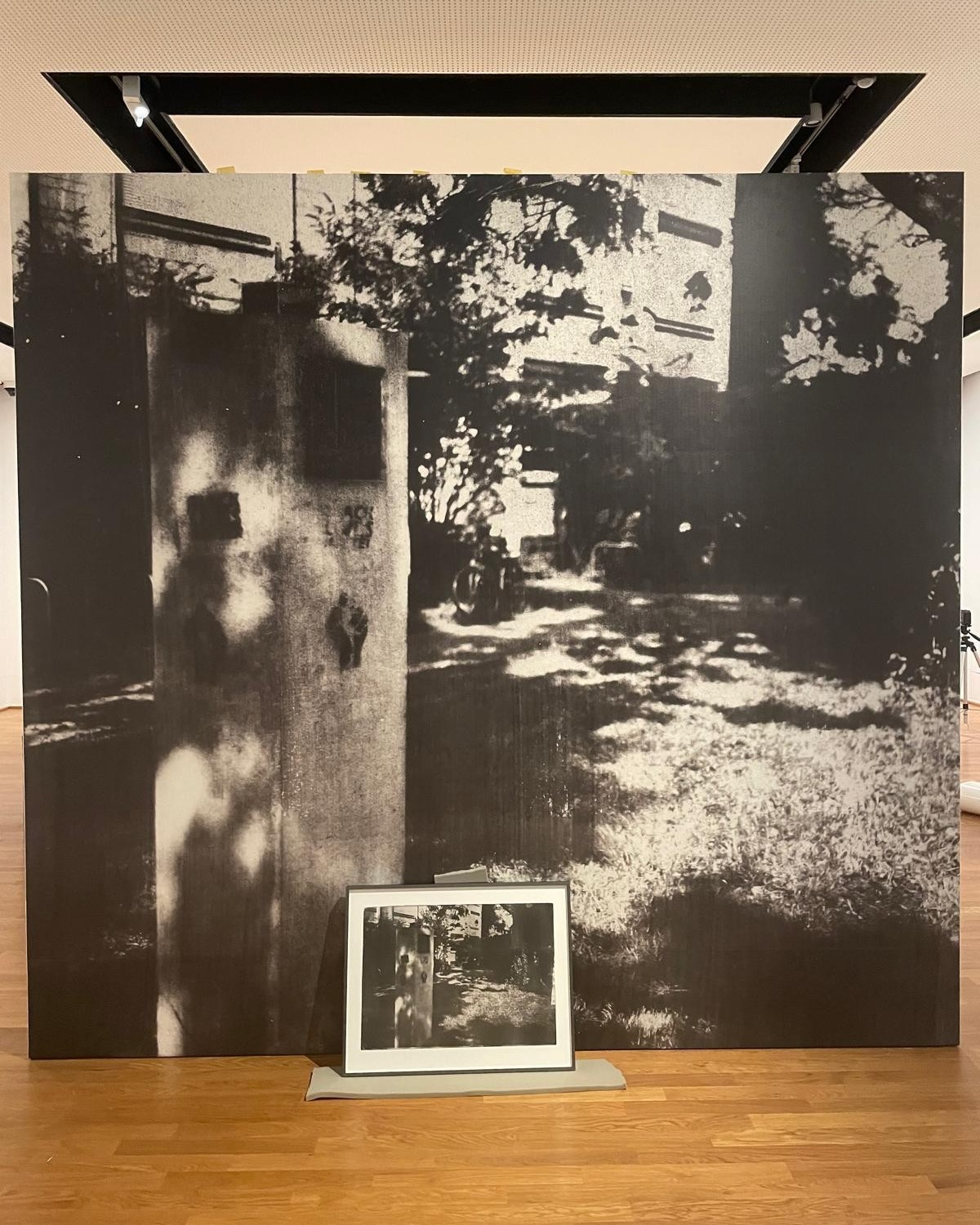 Großformatiges Schwarz-Weiß-Foto einer Straßenszene mit Gebäude, Bäumen und Schattenwurf, davor auf einem Podest ein kleineres gerahmtes Foto derselben Szene. (KI-Alternativtext)