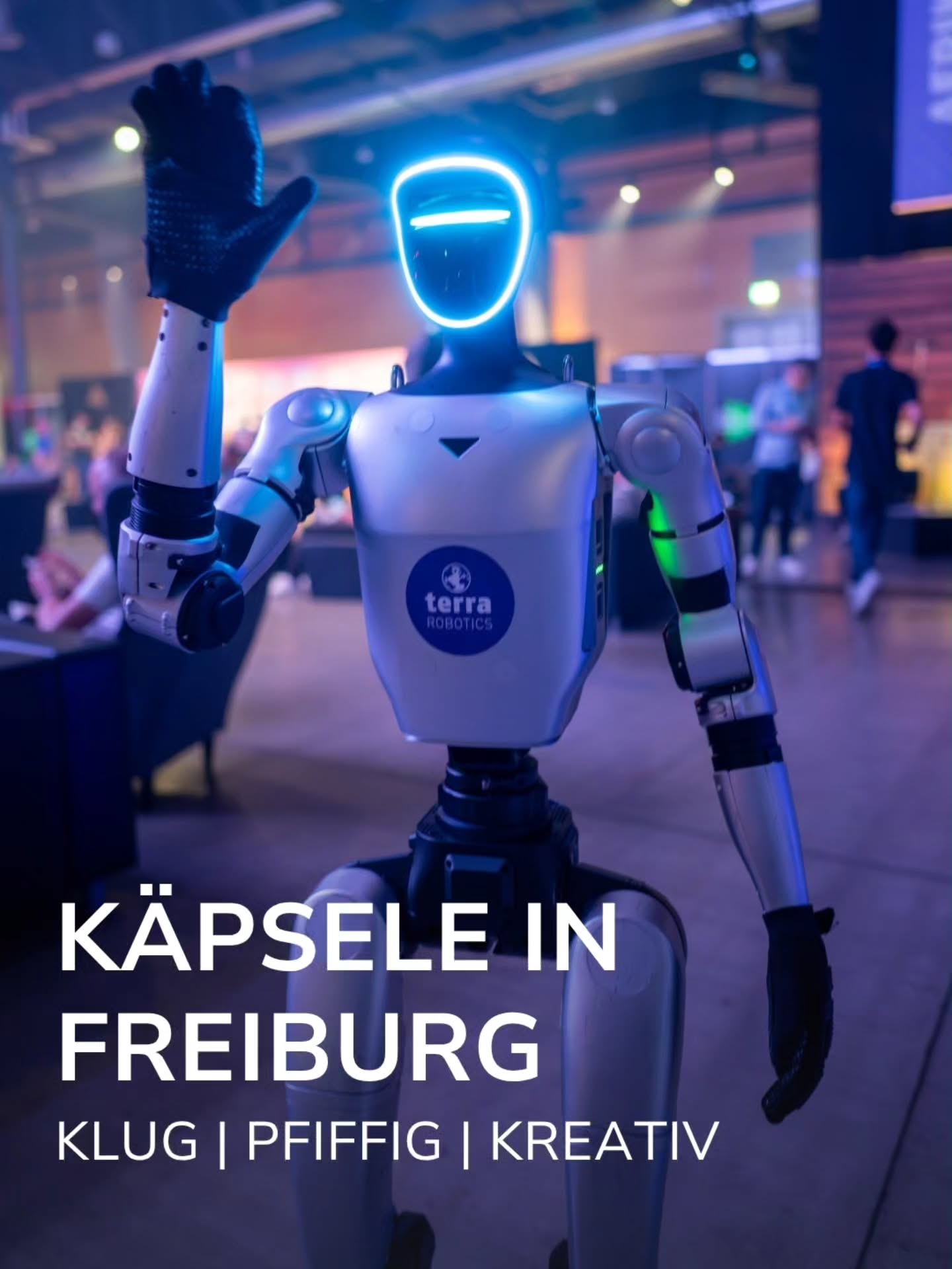 Ein weißer humanoider Roboter mit leuchtend blauem Kopfring winkt mit der rechten Hand. Auf der Brust ist das Logo "terra ROBOTICS" zu sehen. Im unteren Bildbereich steht der weiße Text "KÄPSELE IN FREIBURG" sowie darunter "KLUG | PFIFFIG | KREATIV". Der Hintergrund zeigt einen unscharfen Veranstaltungsort mit Personen. (KI-Alternativtext)