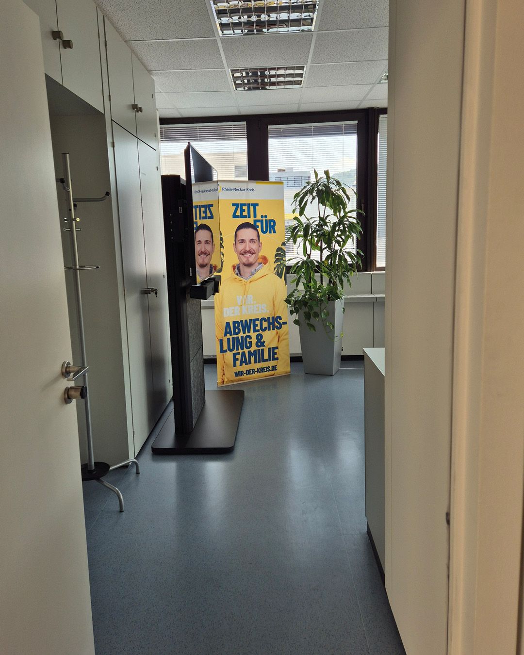 Blick durch eine Türöffnung in einen hellen Büroflur mit weißen Schränken links und einer großen Zimmerpflanze rechts. Im Zentrum steht ein gelbes Standbanner mit dem Text "ZEIT FÜR ABWECHSLUNG & FAMILIE" und einem Bild einer lächelnden Person in einem gelben Hoodie. (KI-Alternativtext)