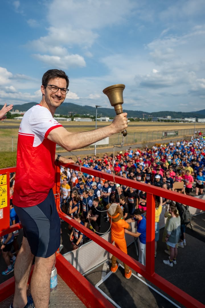 Eine Person in einem rot-weißen T-Shirt steht auf einer roten Plattform und hält eine große Glocke in die Höhe. Im Hintergrund versammelt sich eine große Menschenmenge in Sportkleidung auf einer Wiese unter blauem Himmel. (KI-Alternativtext)