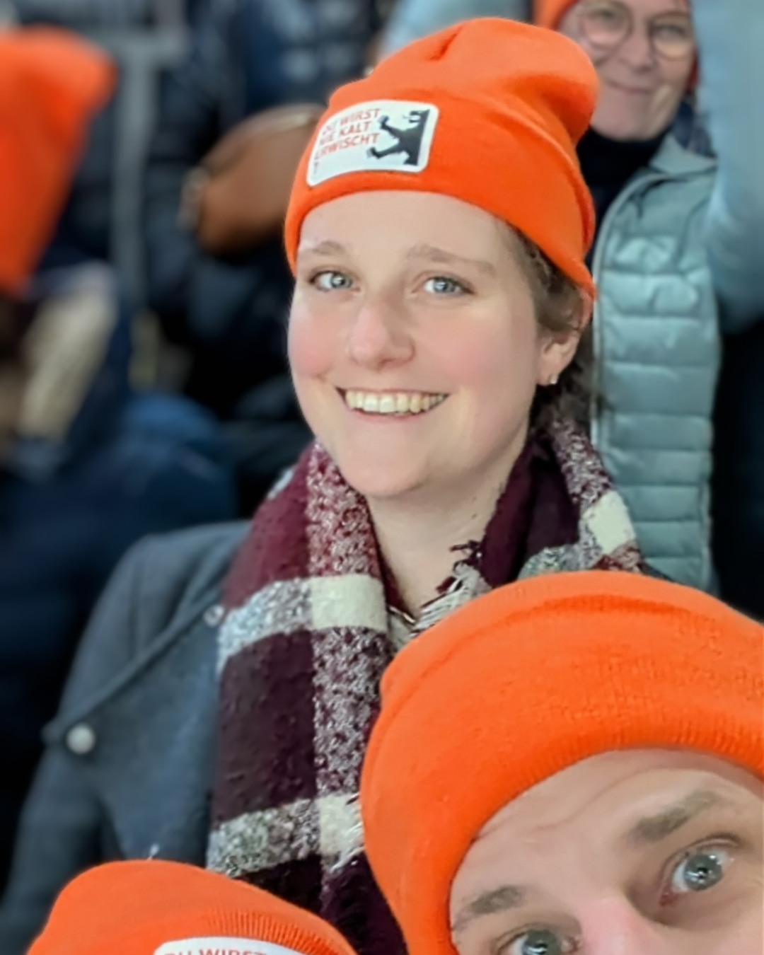 Dieser Alternativtext wurde KI-generiert: Ein Porträt einer lächelnden Person mit einer roten Mütze. Im Hintergrund sind weitere Personen mit roten Mützen zu sehen.