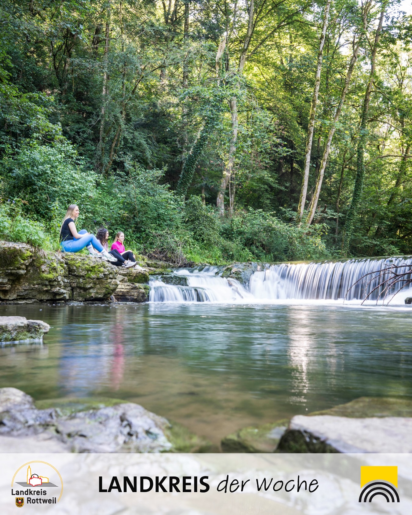 Personen sitzen am Ufer eines Flusses mit einem kleinen Wasserfall im Hintergrund. Um den Fluss ist ein Wald zu sehen.