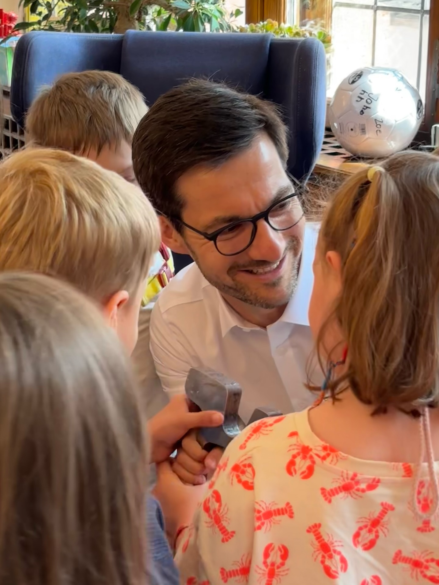 Eine Person mit Brille und weißem Hemd lächelt und blickt auf eine Gruppe von Kindern, von denen eines einen grauen Gegenstand hält. Im Hintergrund steht ein silberner Ballon mit den Aufschriften „Joha“ und „Zoe“. (KI-Alternativtext)