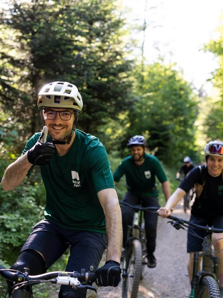 Dieser Alternativtext wurde KI-generiert: Eine Gruppe von Personen fährt auf Fahrrädern einen Waldweg entlang. Die vorderste Person zeigt mit dem Finger nach vorne und lächelt.