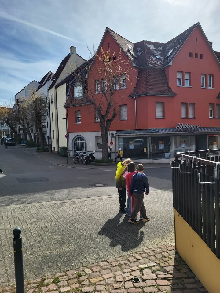 Dieser Alternativtext wurde KI-generiert: Ein Straßenbild zeigt mehrere Gebäude mit roten Fassaden und ein paar Bäume. Im Vordergrund stehen einige Personen auf einem Bürgersteig.