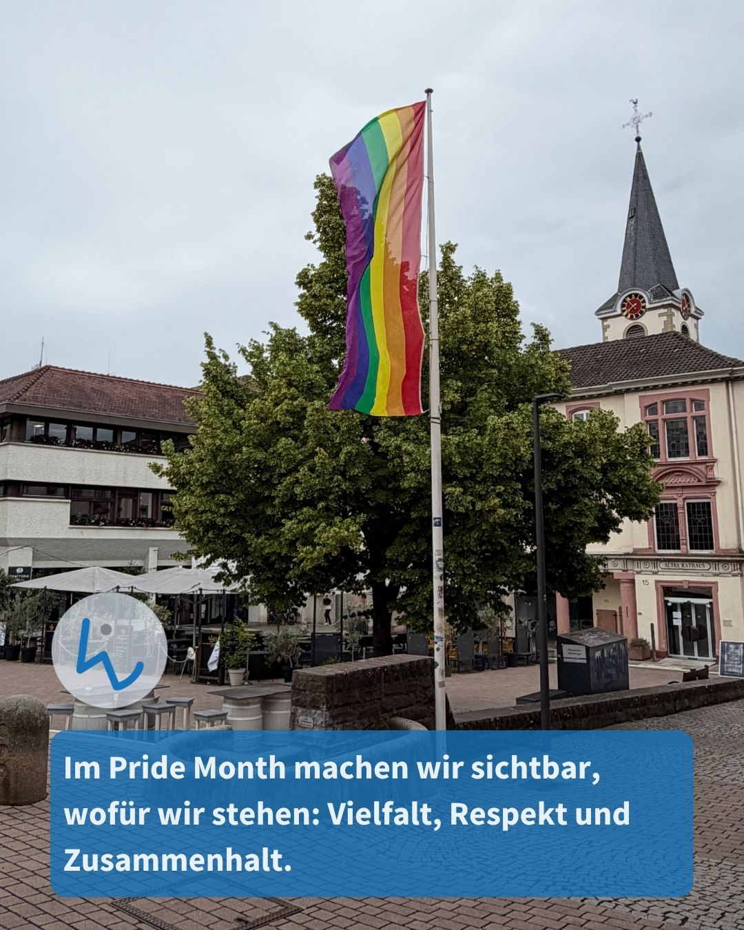 Eine Regenbogenflagge weht an einem Mast auf einem Platz vor einem Rathaus mit Turm. Ein blauer Textkasten unten im Bild verkündet: „Im Pride Month machen wir sichtbar, wofür wir stehen: Vielfalt, Respekt und Zusammenhalt.“ (KI-Alternativtext)