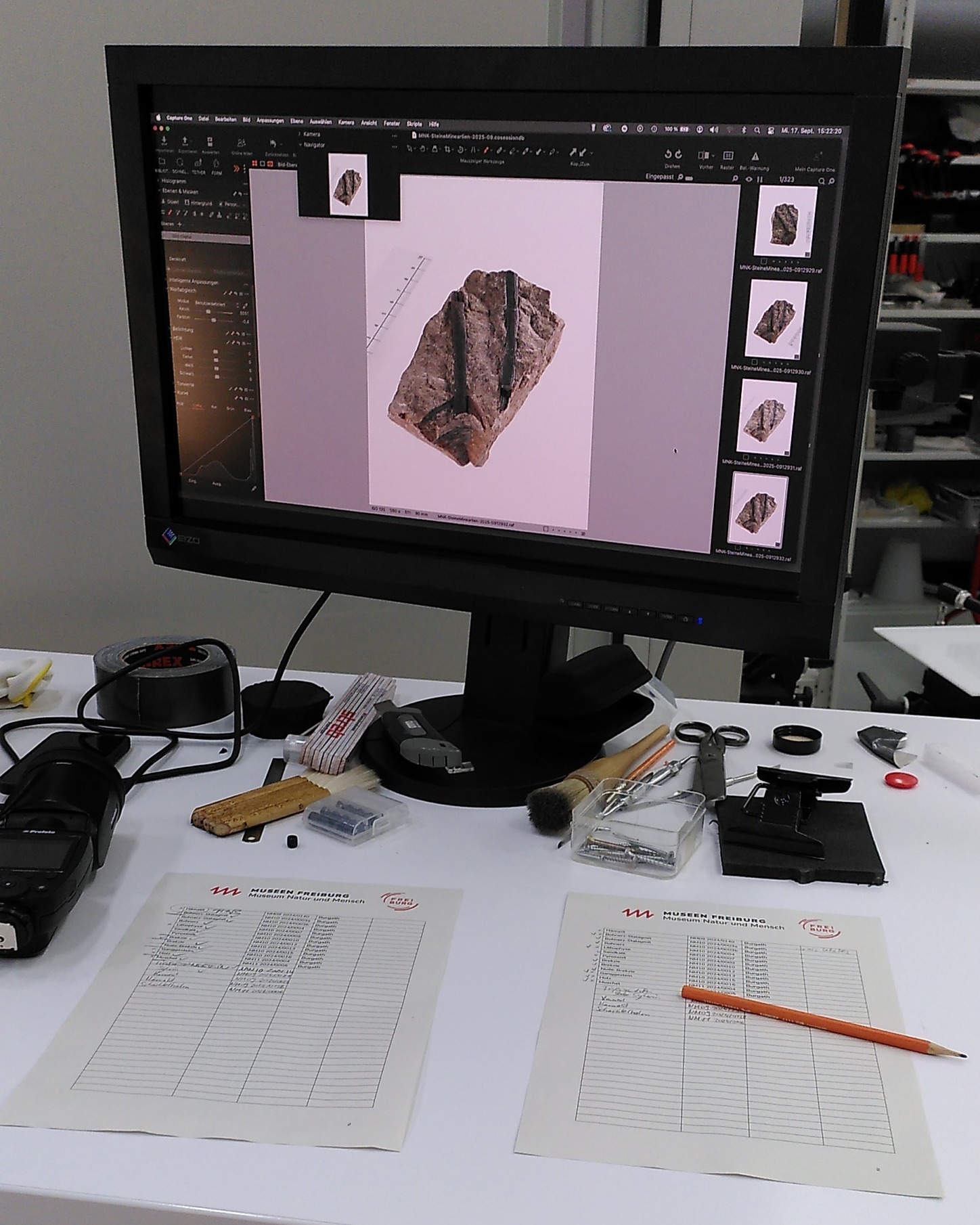 Dieser Alternativtext wurde KI-generiert: Ein Computerbildschirm zeigt detaillierte Aufnahmen eines Gesteinsstücks. Auf dem Tisch davor befinden sich Arbeitsmaterialien, Dokumente und Werkzeuge.