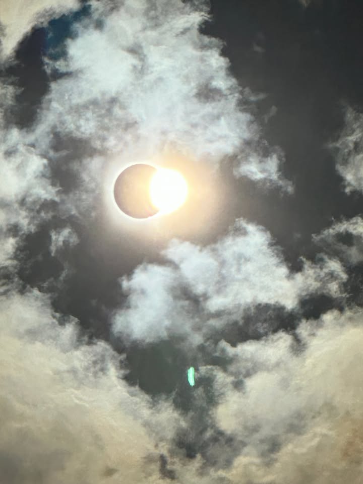 Die Sonne wird während einer partiellen Sonnenfinsternis teilweise vom Mond verdeckt und leuchtet hell durch eine Lücke in den Wolken. Umgebende weiße und graue Wolken rahmen das Ereignis ein. Ein kleiner grüner Lichtreflex ist unterhalb der Sonne sichtbar. (KI-Alternativtext)