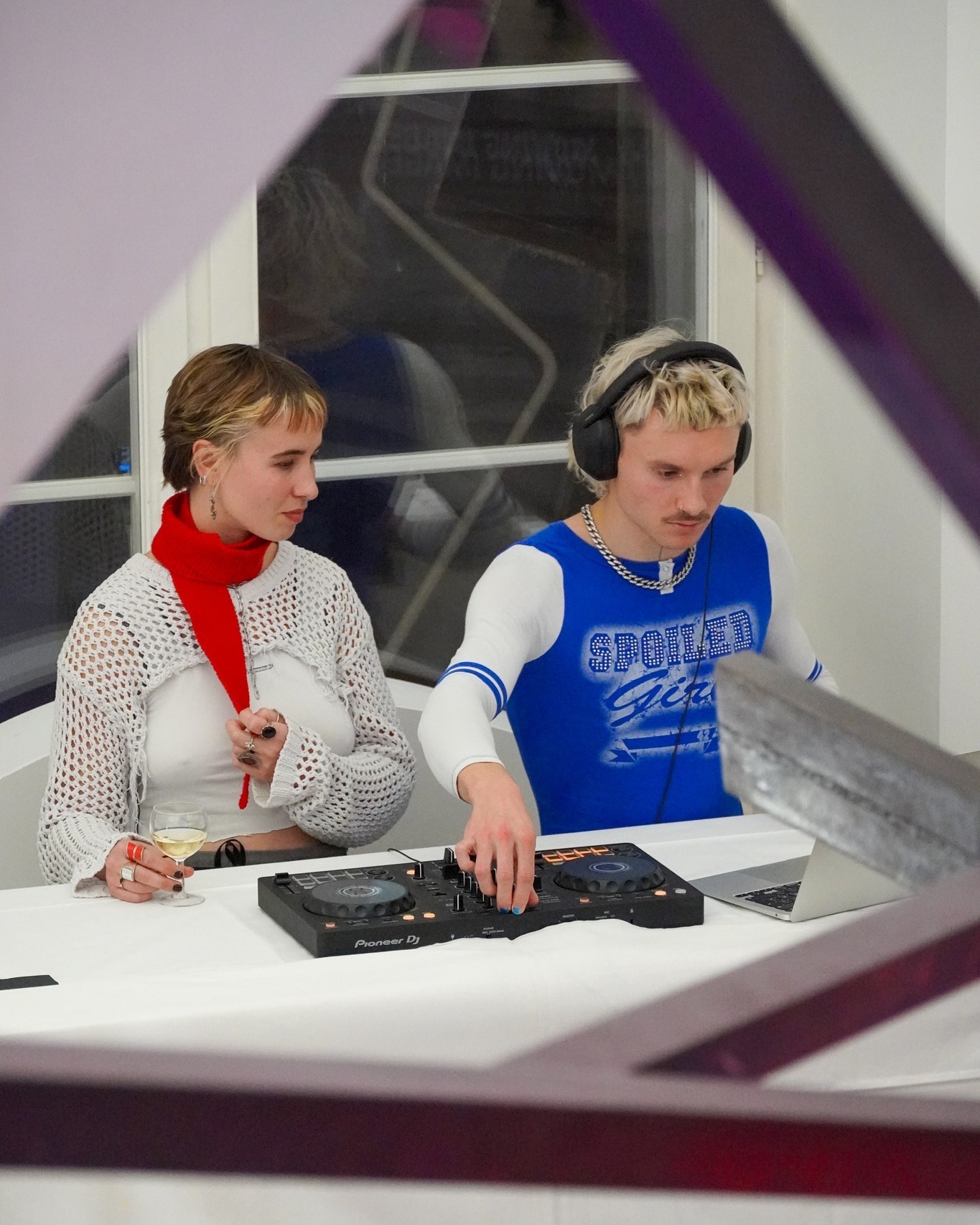 Dieser Alternativtext wurde KI-generiert: Zwei Personen befinden sich in einem Raum mit einer weißen Einrichtung. Eine Person bedient ein Mischpult, während die andere Person daneben sitzt und ein Glas hält.