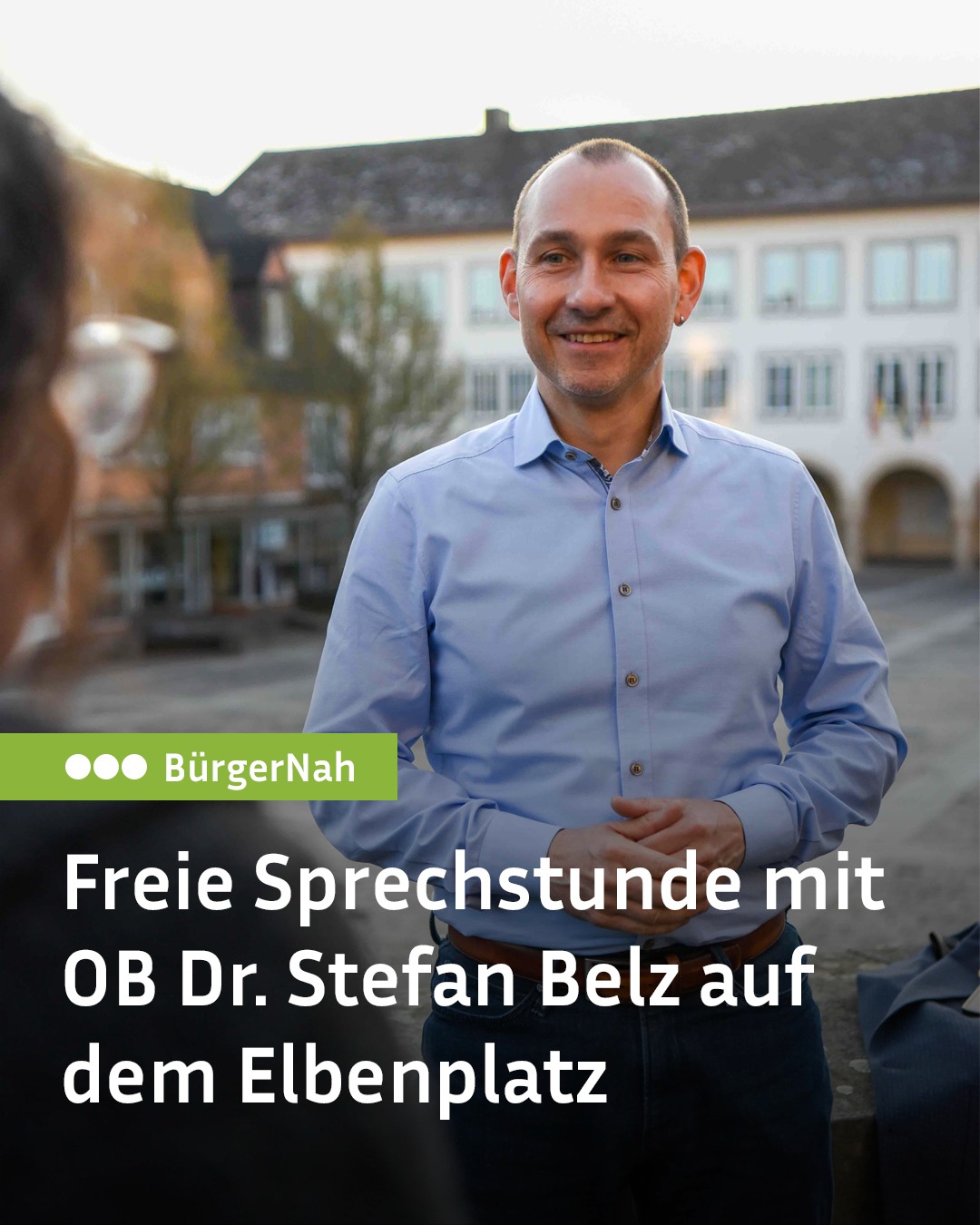 Eine Person in einem hellblauen Hemd steht im Freien und lächelt. Der Text im Bild lautet: „BürgerNah Freie Sprechstunde mit OB Dr. Stefan Belz auf dem Elbenplatz“. (KI-Alternativtext)