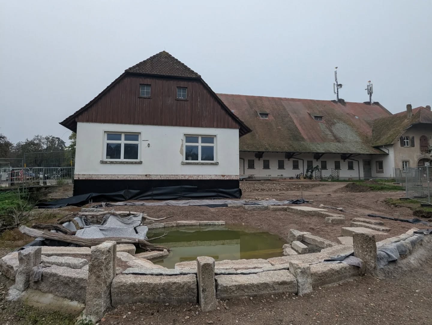 Dieser Alternativtext wurde KI-generiert: Ein Gebäude mit einem roten und einem weißen Teil steht neben einem längeren, niedrigeren Gebäude mit einem grasbewachsenen Dach. Im Vordergrund befindet sich ein Teich, der von Steinen umgeben ist.