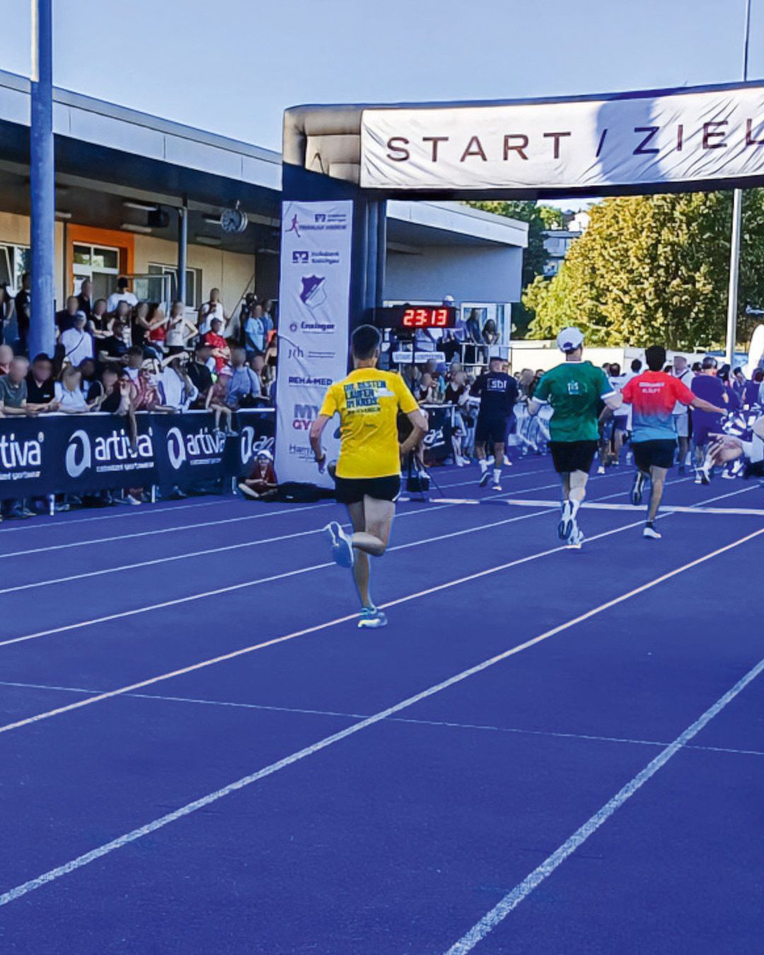 Mehrere Läufer rennen auf einer blauen Laufbahn unter einem Tor mit der Aufschrift START / ZIEL hindurch. Der vorderste Läufer trägt ein gelbes Shirt mit dem Text DIE BESTEN LAUFEN IM KREIS, während eine digitale Uhr die Zeit 23:13 anzeigt. Links am Rand beobachten Zuschauer das Rennen hinter einer Absperrung. (KI-Alternativtext)