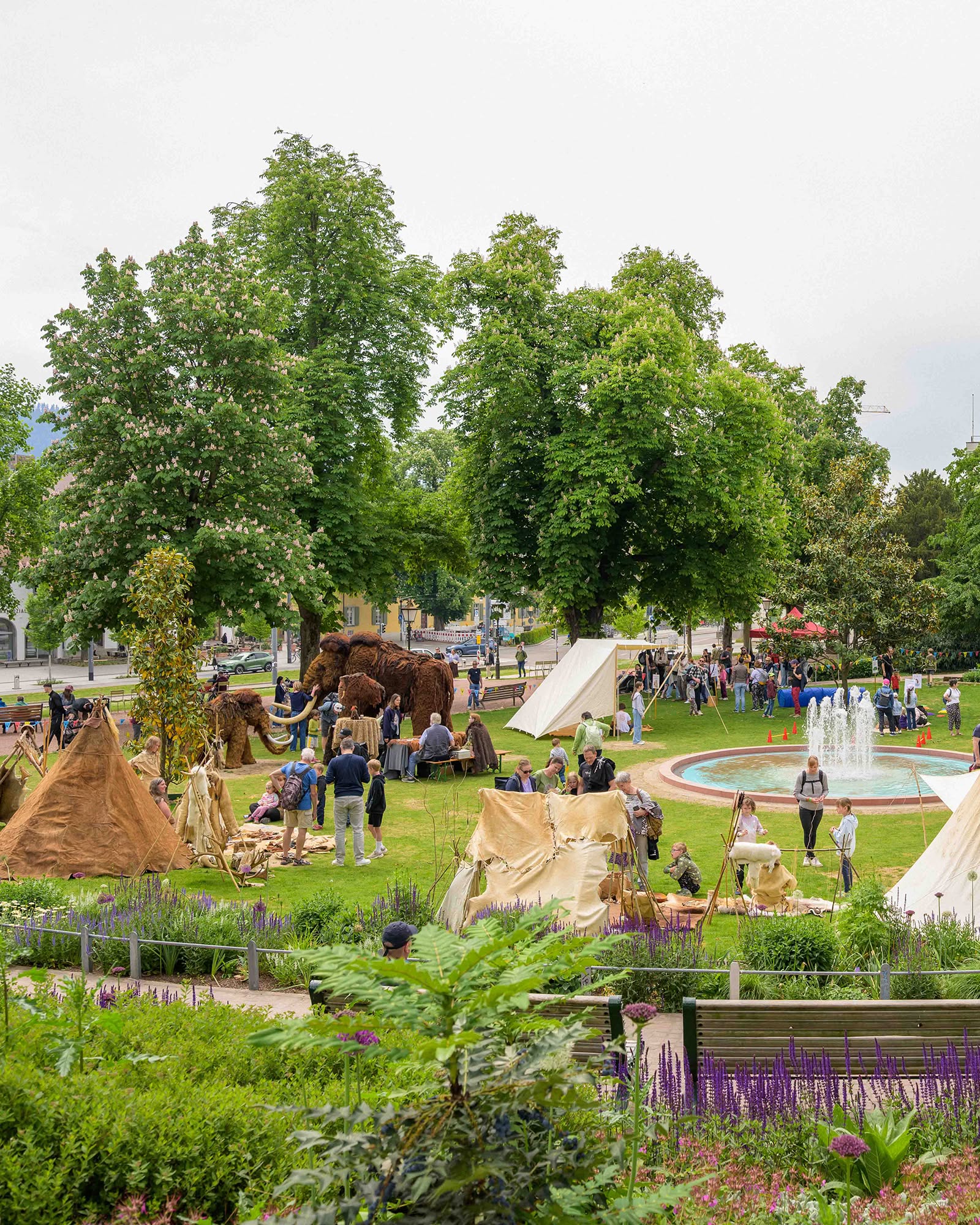 Auf einer Wiese im Park stehen große, behaarte Mammut-Modelle und Zelte aus Stoff und Tierhäuten, umgeben von Menschen, die sich unterhalten oder die Ausstellung betrachten. Im Hintergrund sprudelt ein Brunnen, und hohe Bäume säumen den Platz. (KI-Alternativtext)