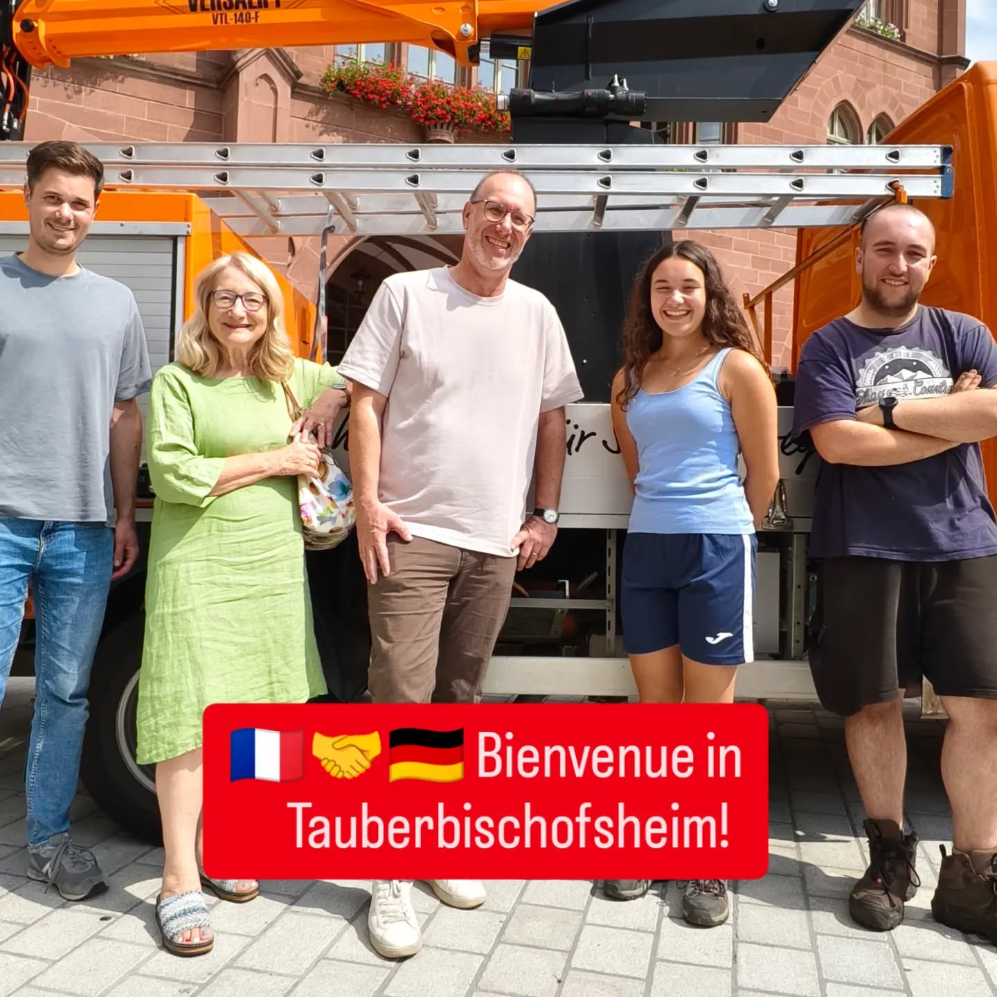 Fünf Personen stehen lächelnd vor einem orangefarbenen Fahrzeug mit Kranarm. Ein roter Balken im Vordergrund zeigt französische und deutsche Flaggen, ein Händedruck-Symbol und den Schriftzug „Bienvenue in Tauberbischofsheim!“. (KI-Alternativtext)